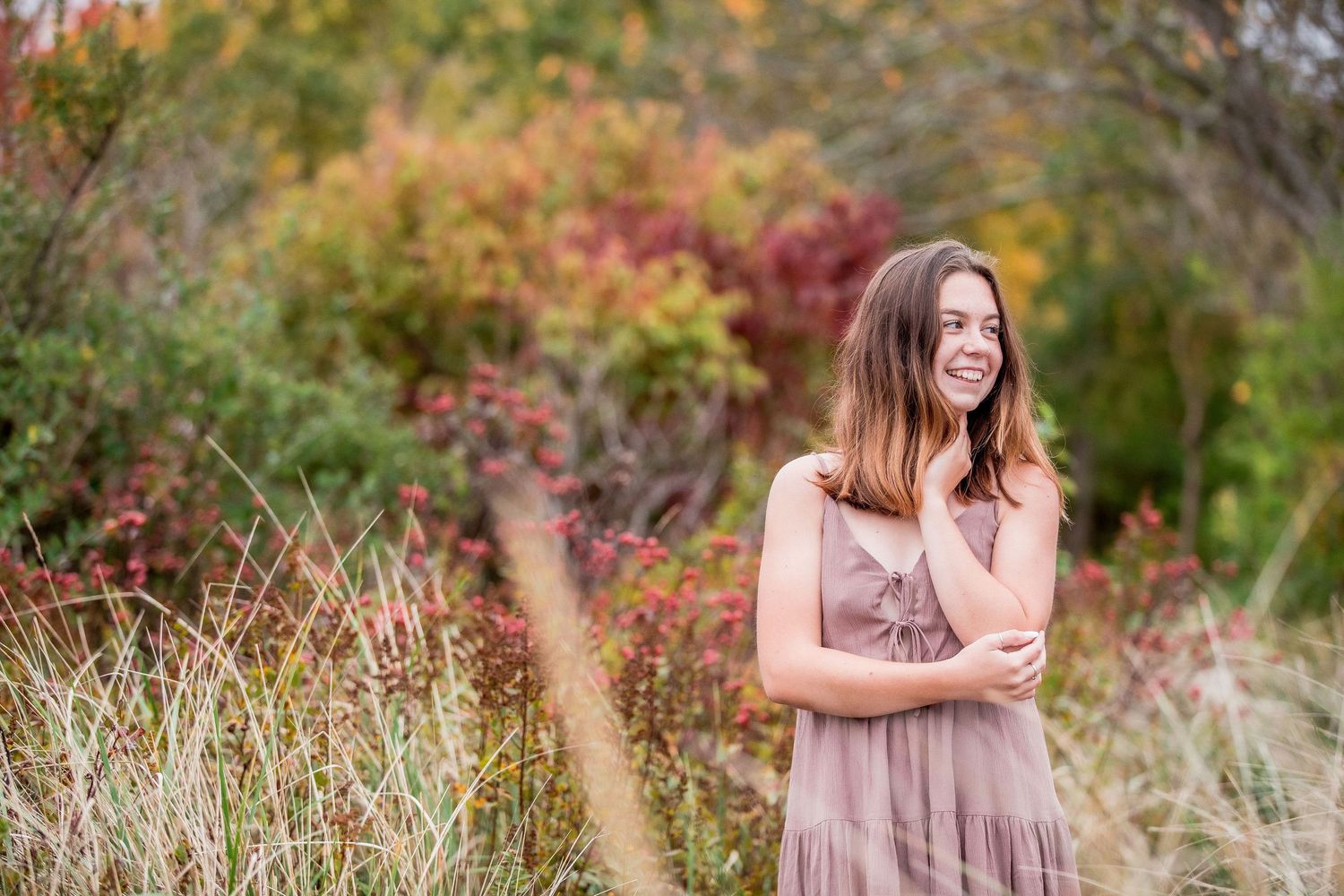 Smiling young woman in mauve dress standing among colorful autumn foliage in a garden.