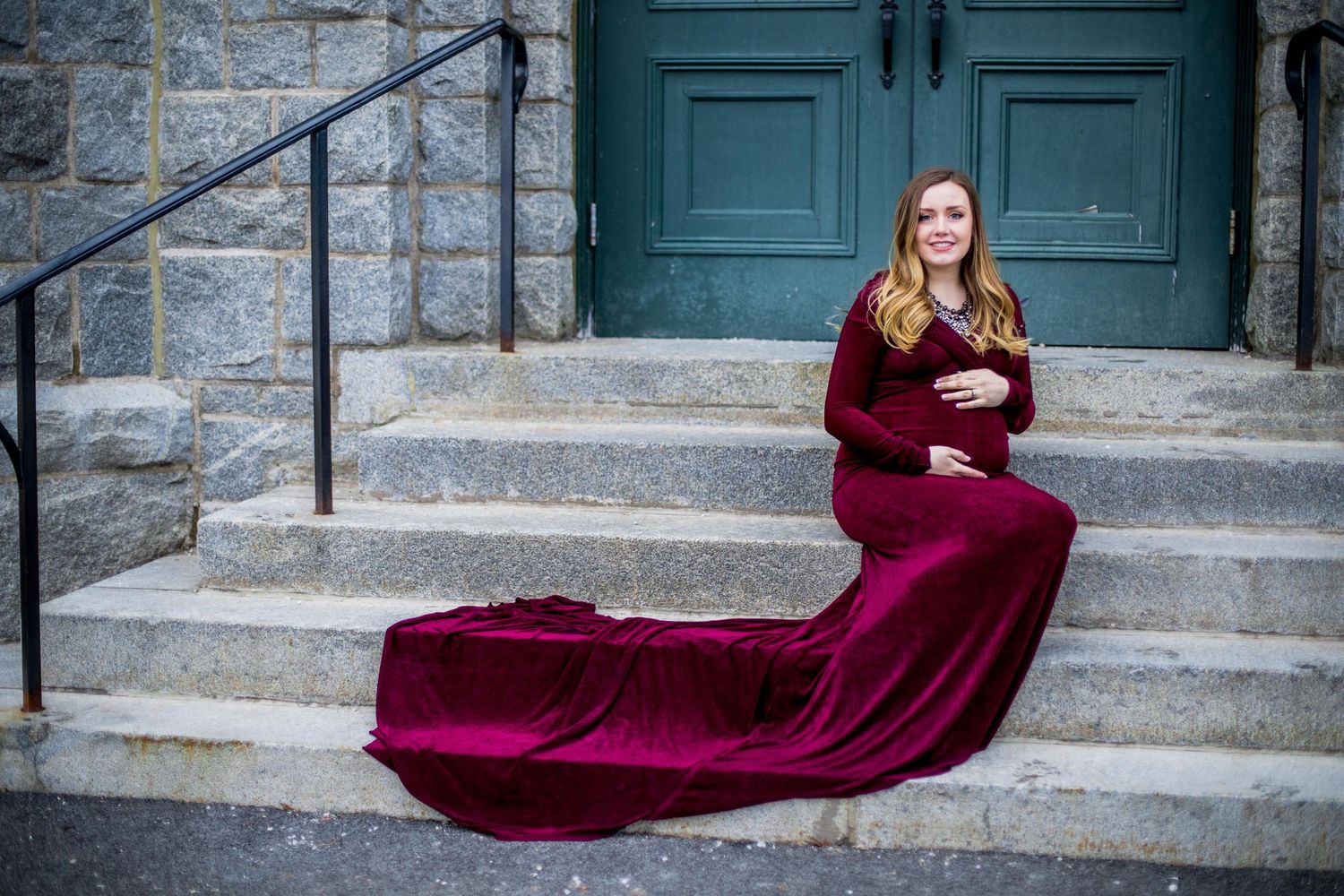 Pregnant woman in burgundy velvet gown with long train sitting gracefully on stone church steps.