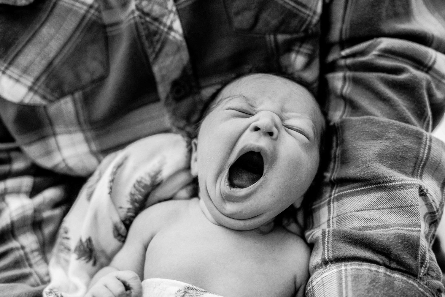 Adorable newborn baby yawning while cradled in parent's arms, captured in black and white photography.