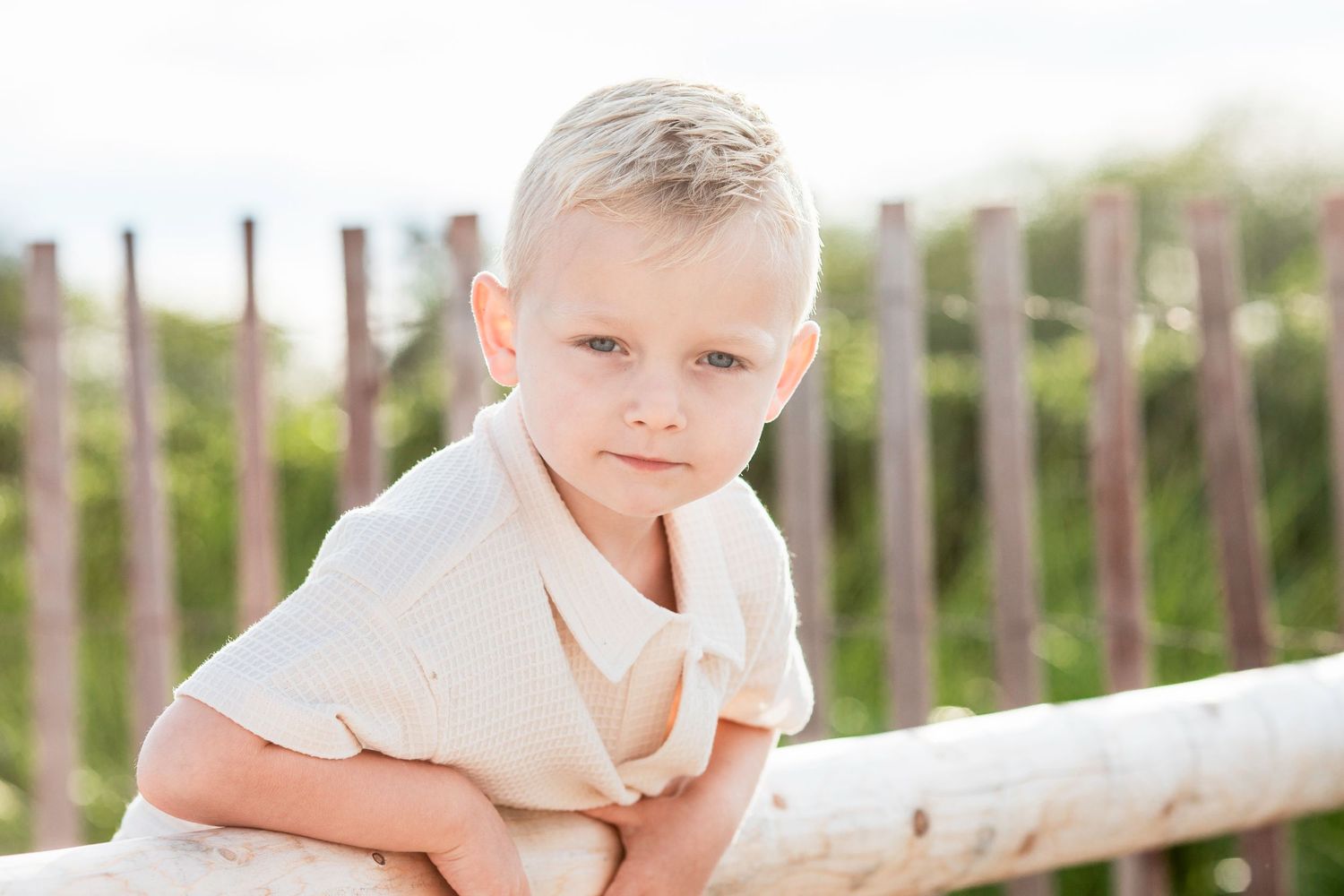 Young blonde boy leaning on a white wooden fence outdoors, wearing a cream shirt with greenery in the background.
