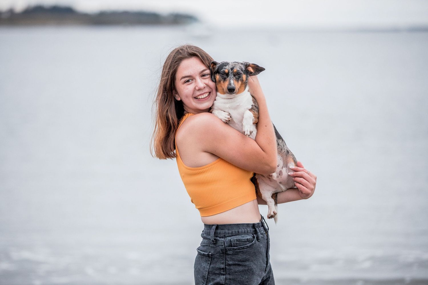 Smiling young woman in orange crop top holding a small dog at the beach.