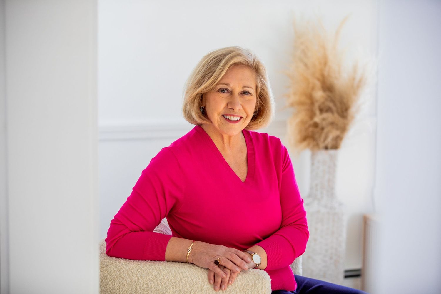 Smiling older woman in pink top seated casually in bright white room with pampas grass decor.