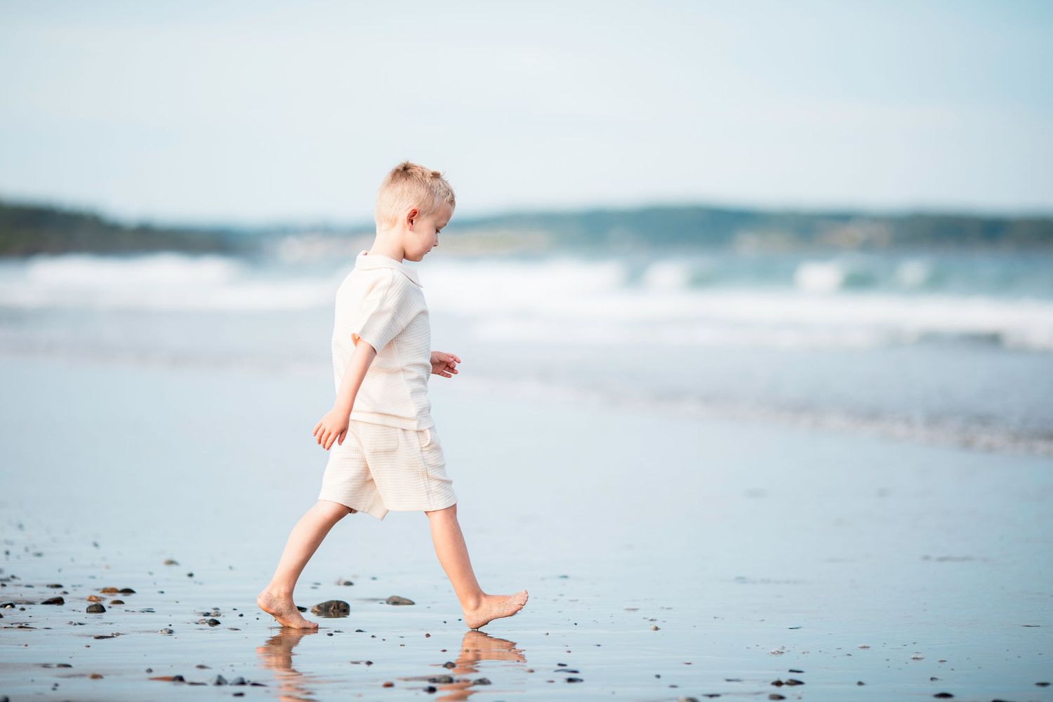 A young blonde child walks barefoot along a serene beach shore in white outfit with ocean waves in background.