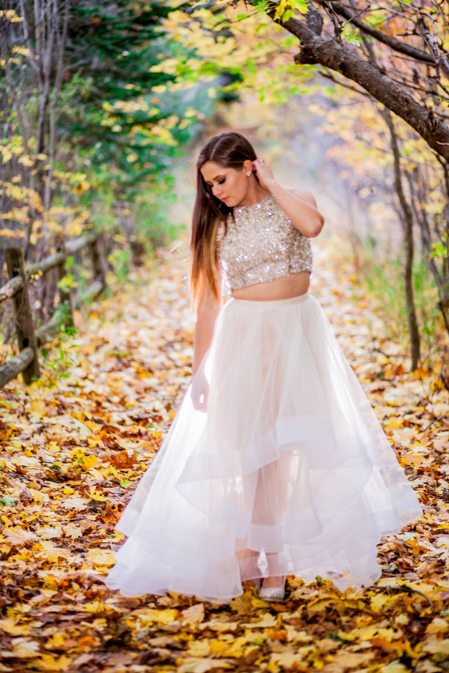 Young woman in white sequin crop top and layered tulle skirt poses on a leaf-covered path in autumn forest.