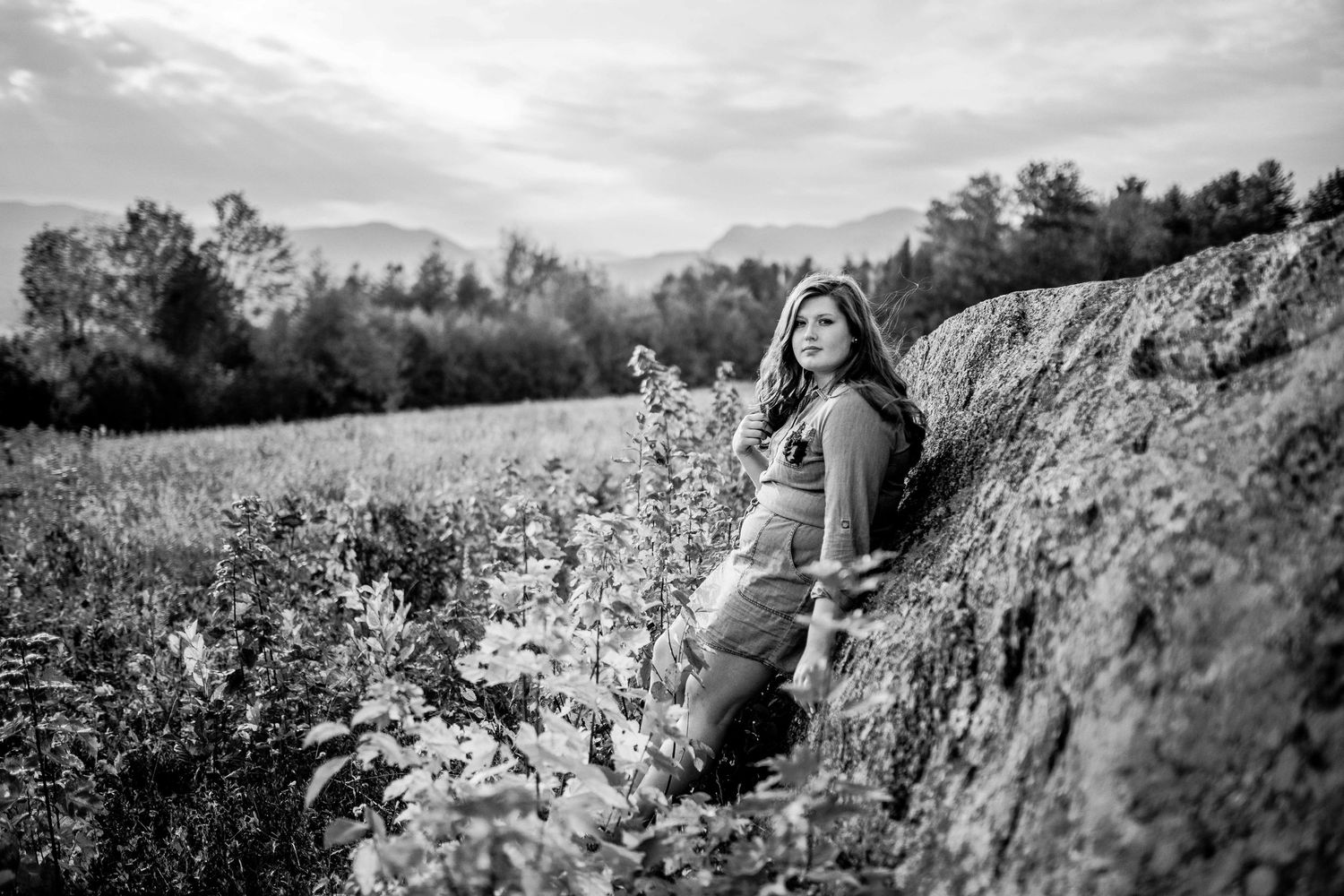 Black and white photo of a young woman leaning against a rocky hillside in a field.