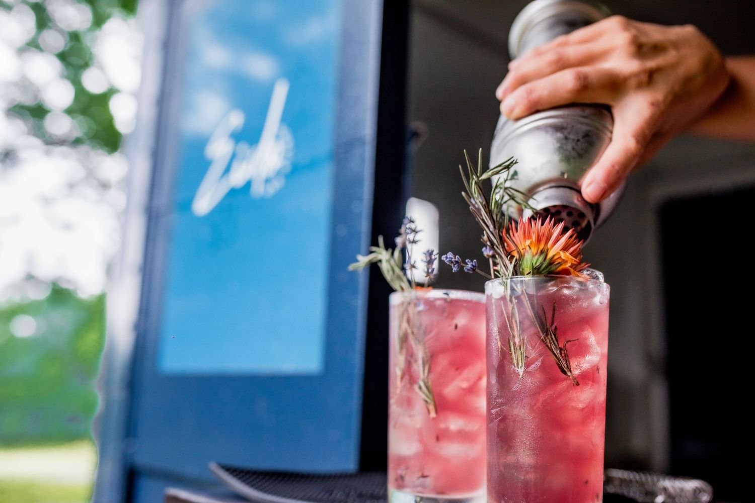 Bartender pouring drink into pink cocktail garnished with rosemary and flower near blue truck.