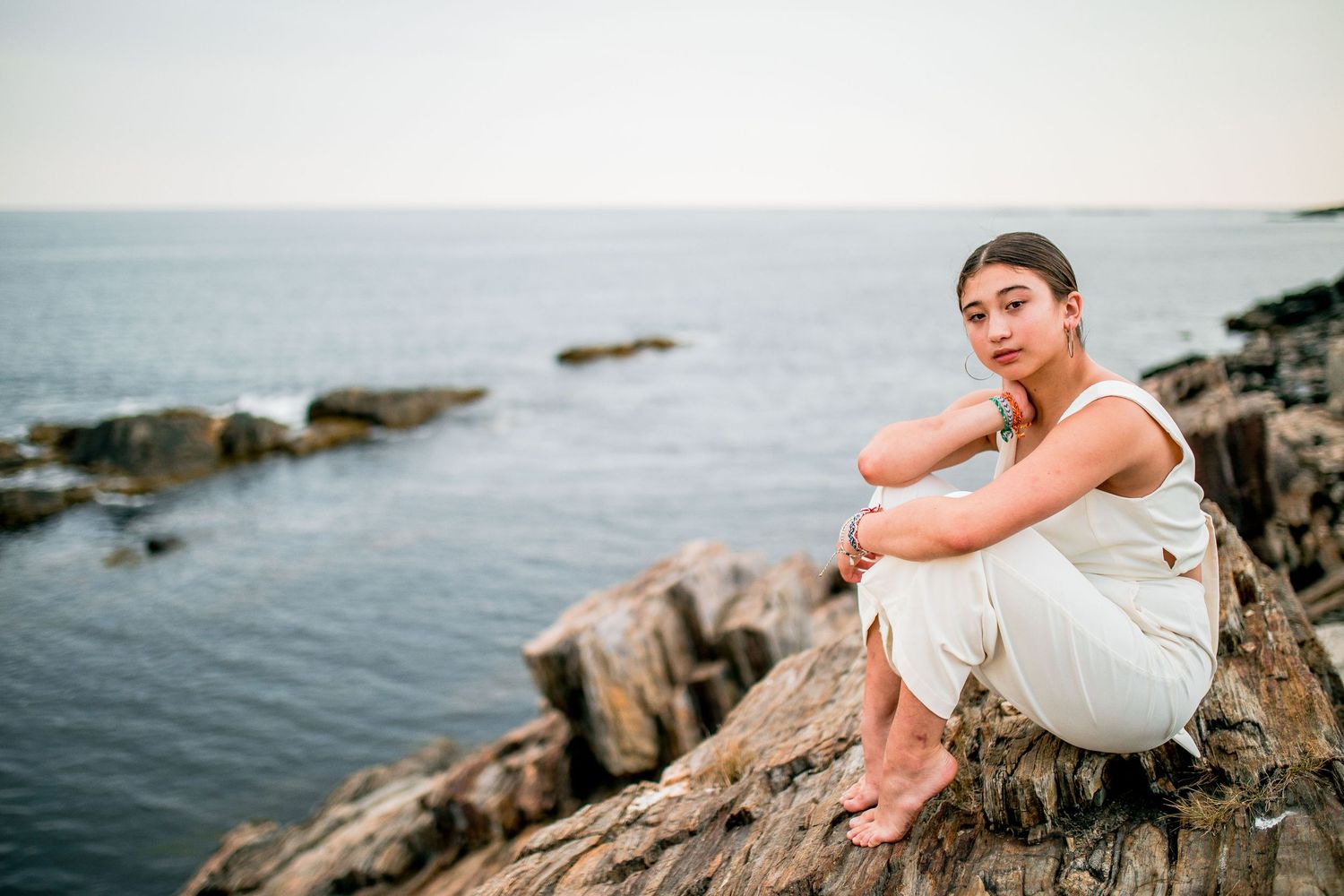 Young woman in white outfit sitting on rocky coastal cliffs overlooking calm ocean at dusk.