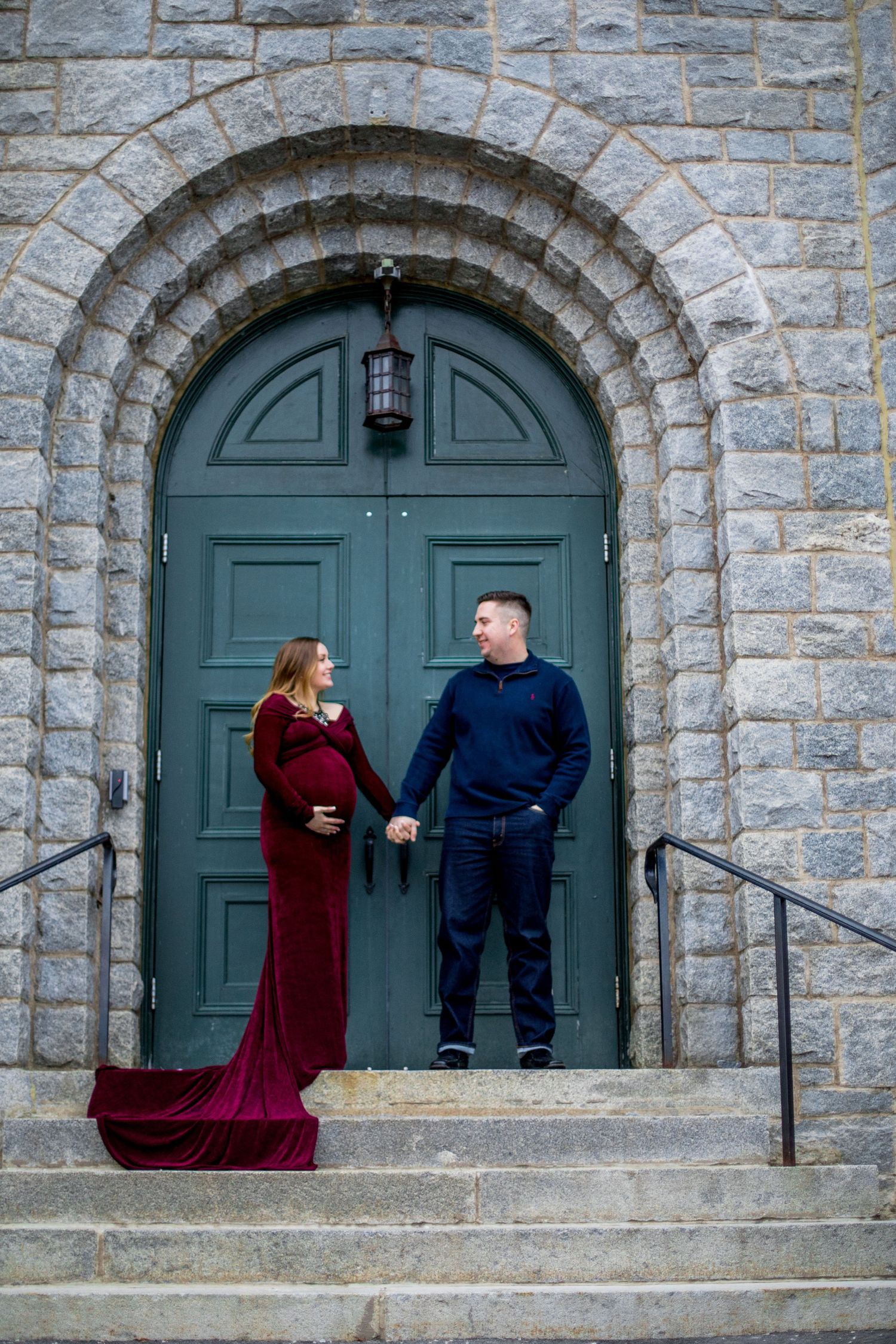 Expecting couple holding hands on stone church steps before green arched door during maternity photo session.