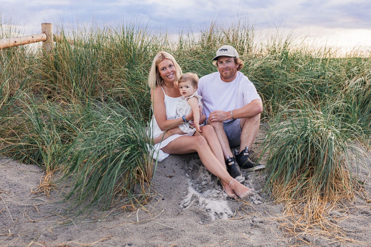 Young family with toddler sitting together in beach grass dunes at sunset.