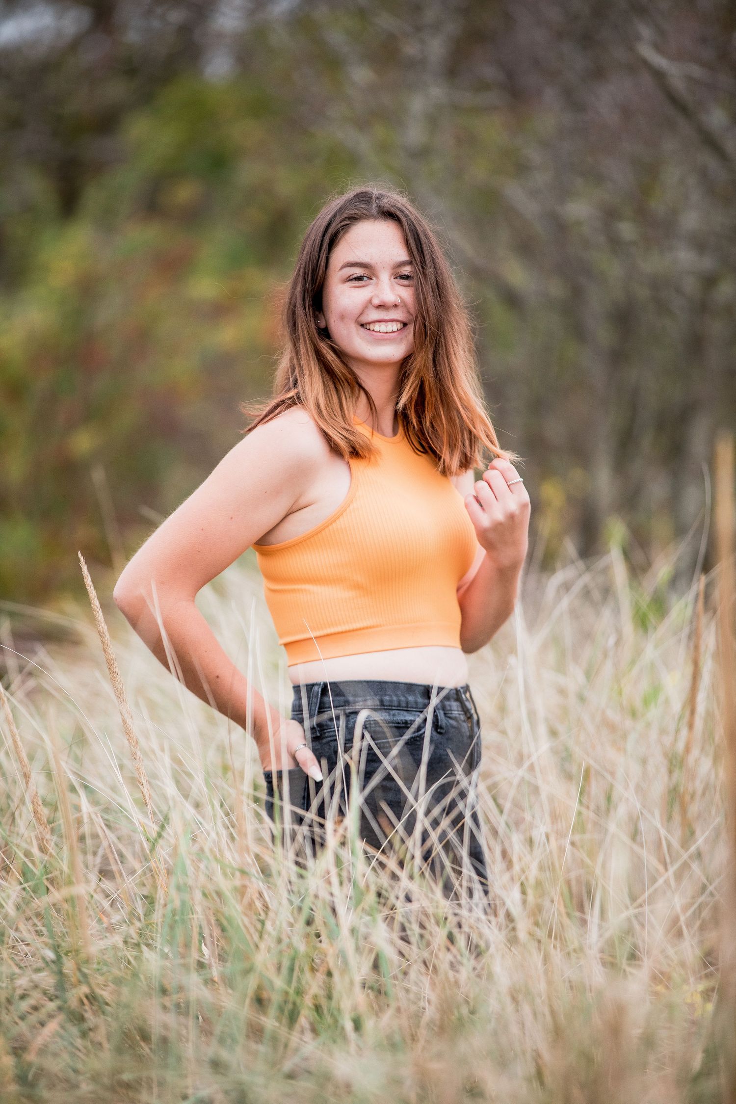 Smiling teenage girl in orange crop top standing in tall golden grass with hand on hip.
