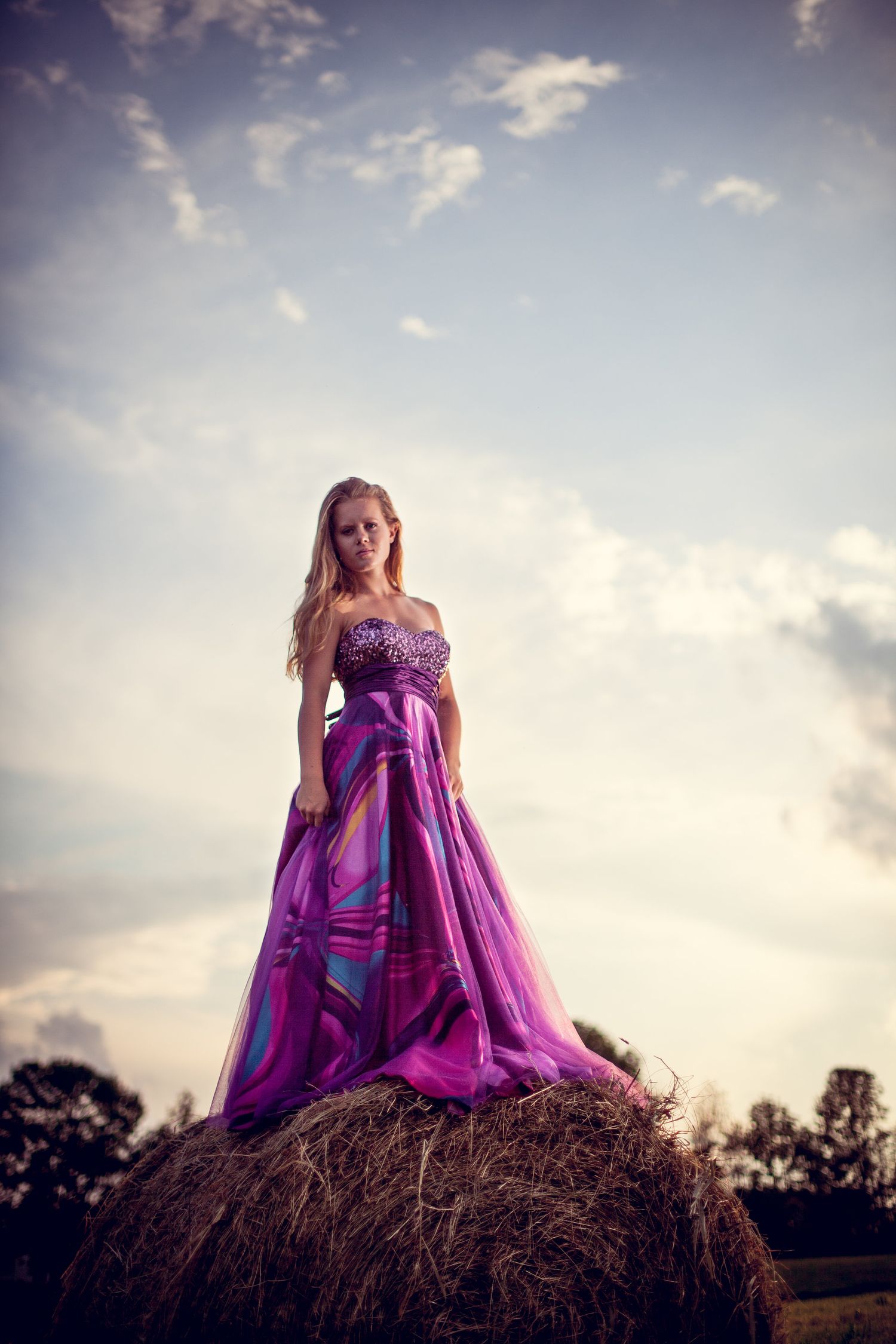 Elegant blonde woman in a purple strapless gown standing atop a hay bale at sunset.
