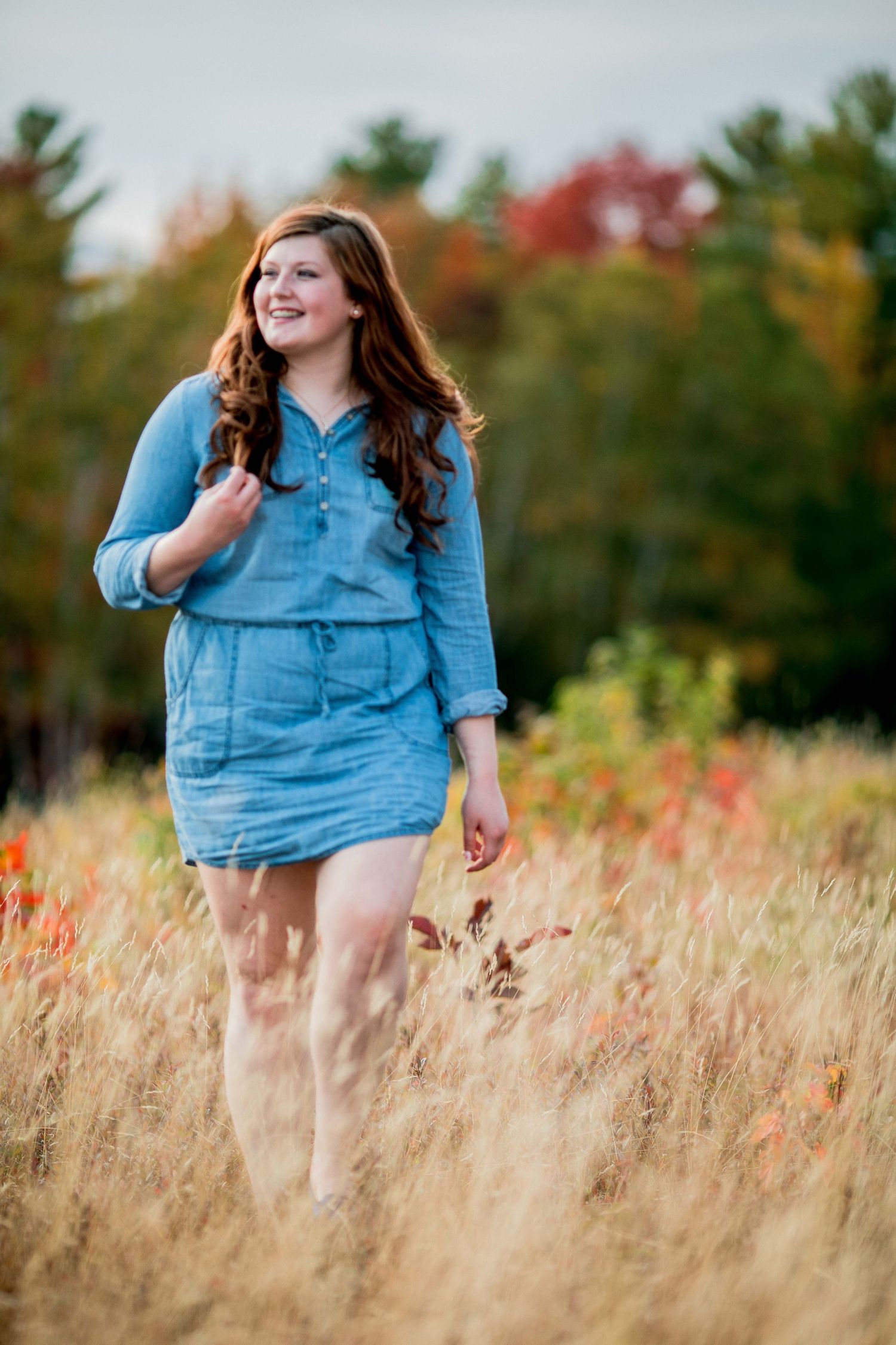 Smiling young woman in blue denim dress walking through golden autumn field with fall foliage.
