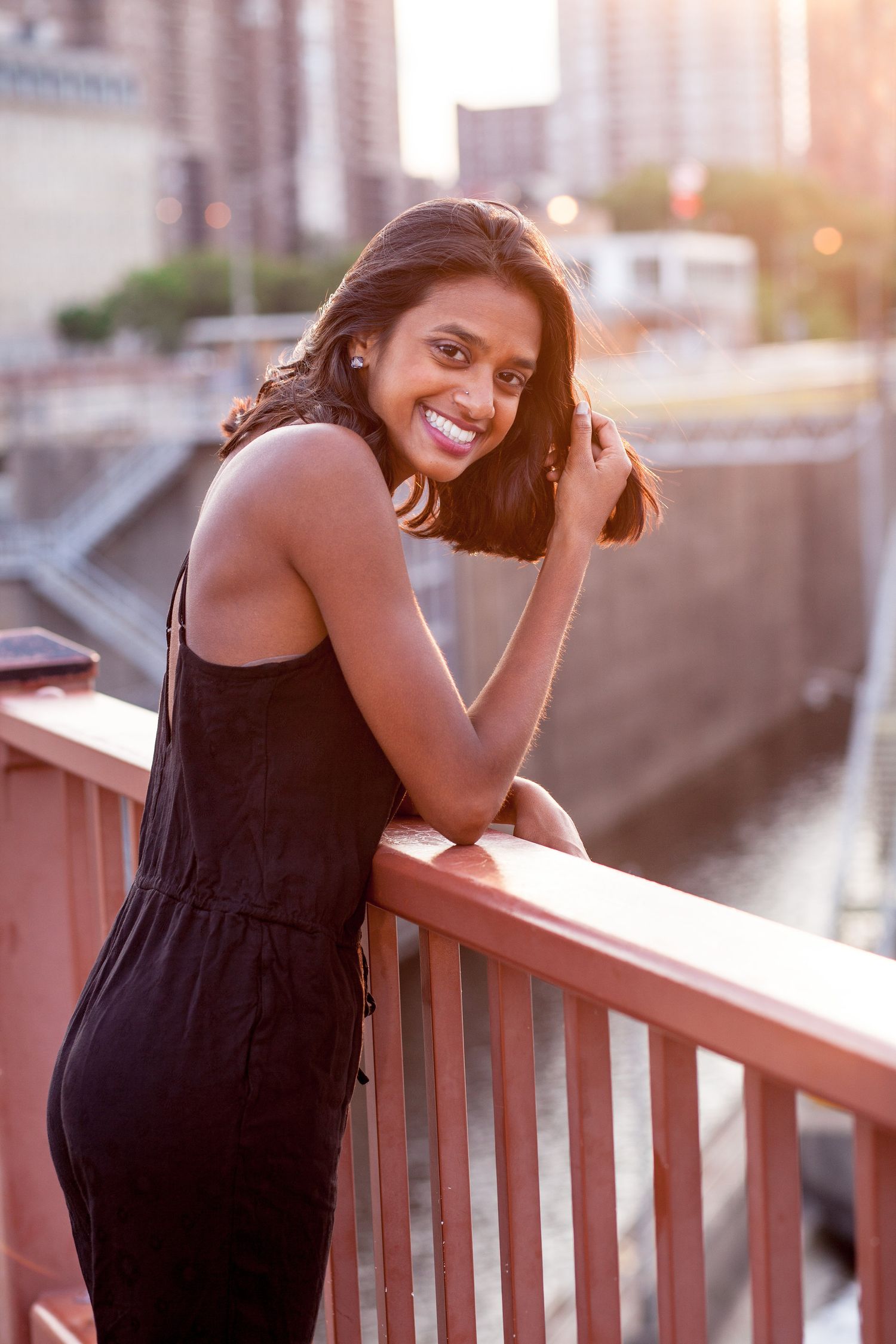 Smiling South Asian woman in black jumpsuit leans on bridge railing with city skyline at sunset.