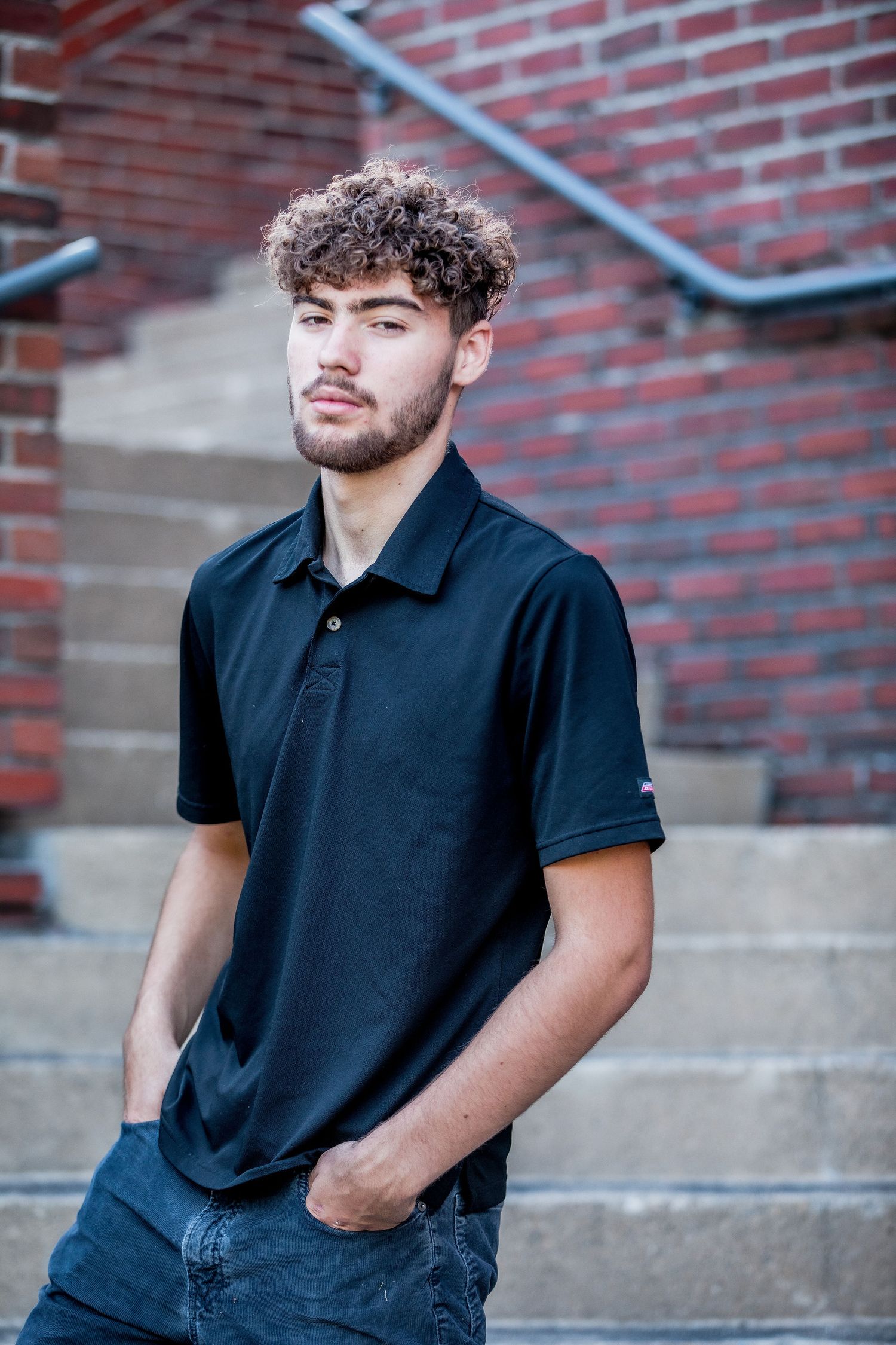 Young man with curly hair in a black polo shirt standing near brick building stairs.