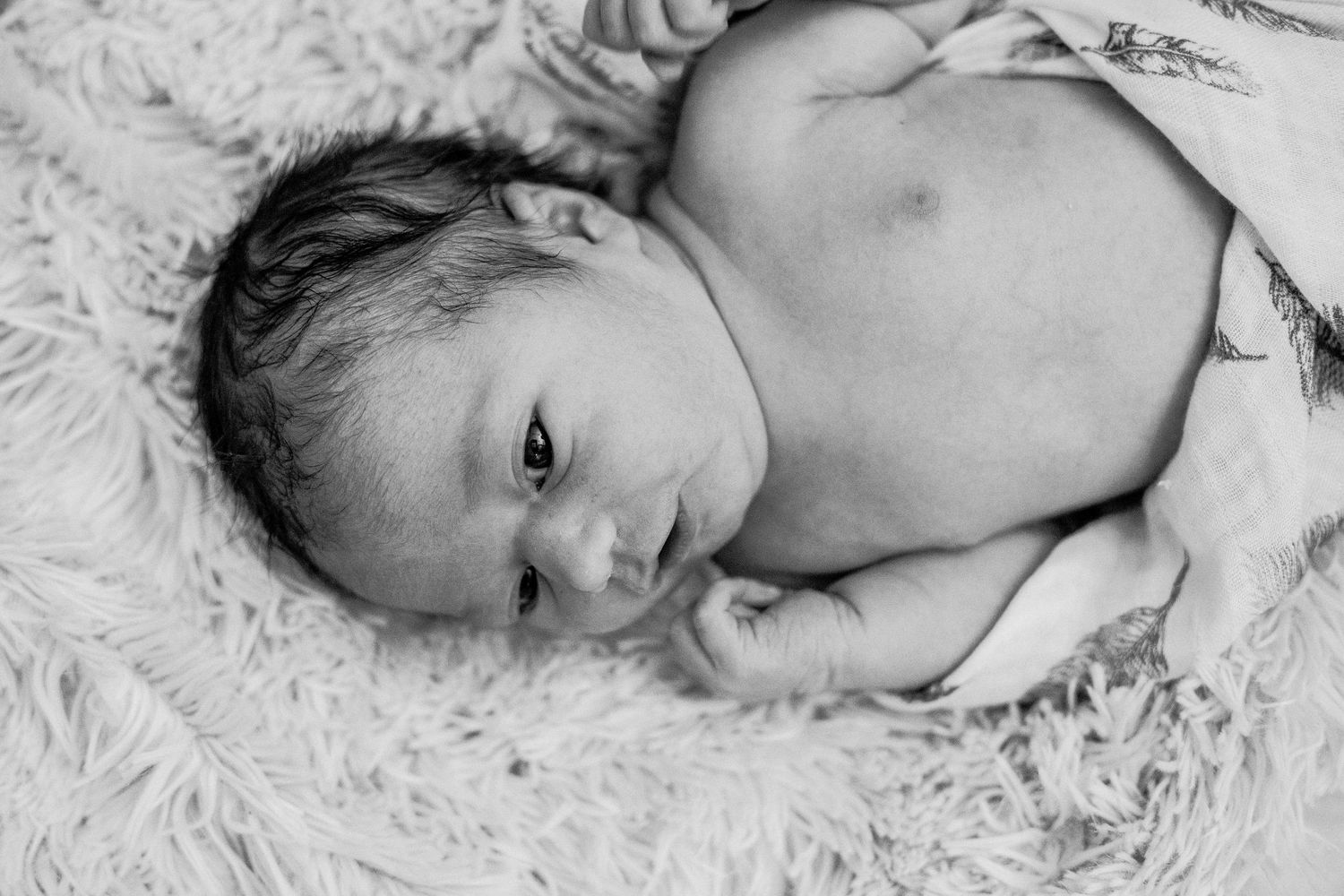 Black and white portrait of a newborn baby lying on a fluffy blanket with eyes open.