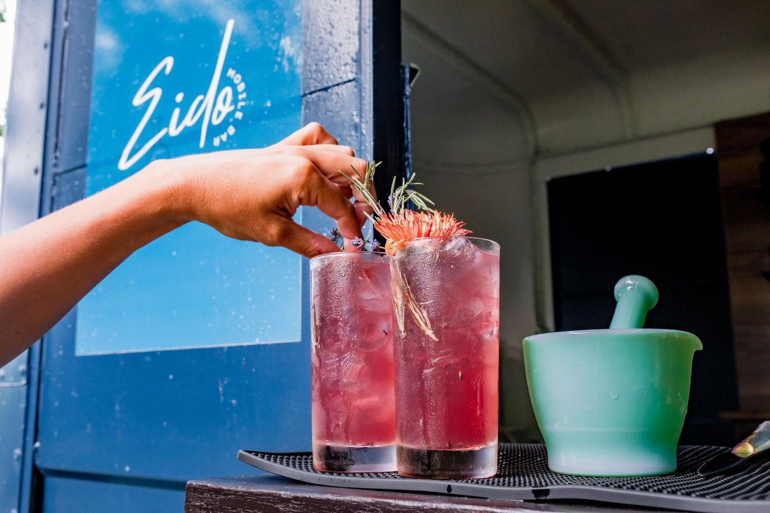 Hand reaching for pink cocktail garnished with rosemary beside blue food truck window.