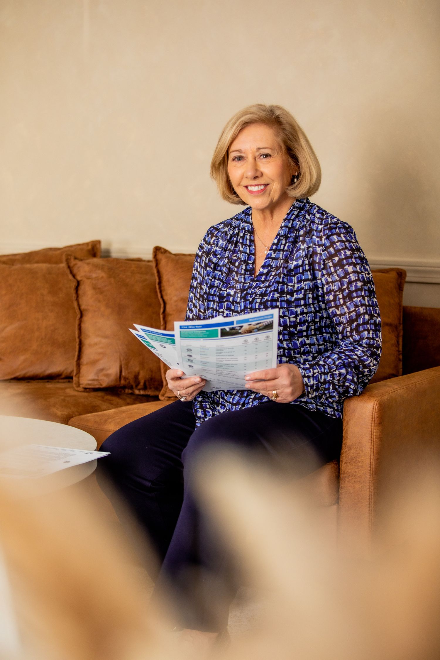 Smiling older woman in a blue patterned blouse reading a magazine on a brown leather sofa.