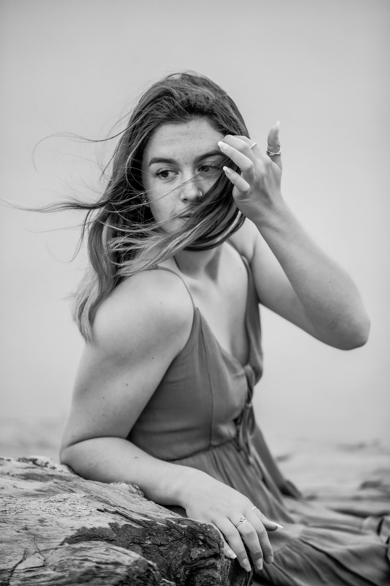 Black and white portrait of a woman brushing hair from her face while sitting on rocks outdoors.