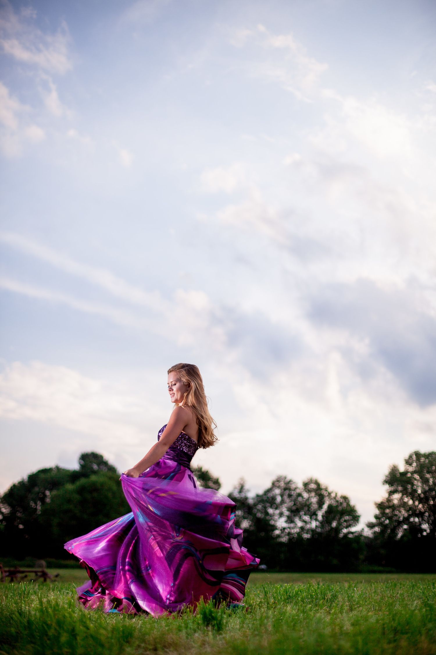 Young woman in a flowing purple gown twirling gracefully in a green field at dusk.