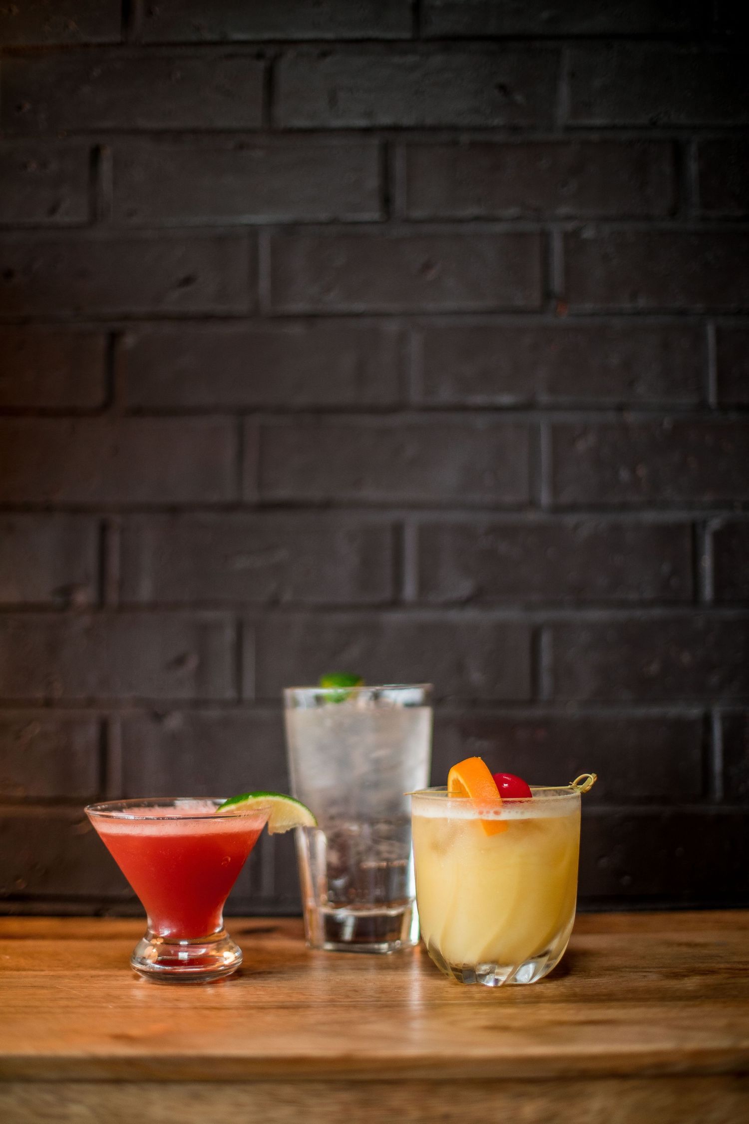 Three colorful cocktails on a wooden bar top against a dark brick wall background.