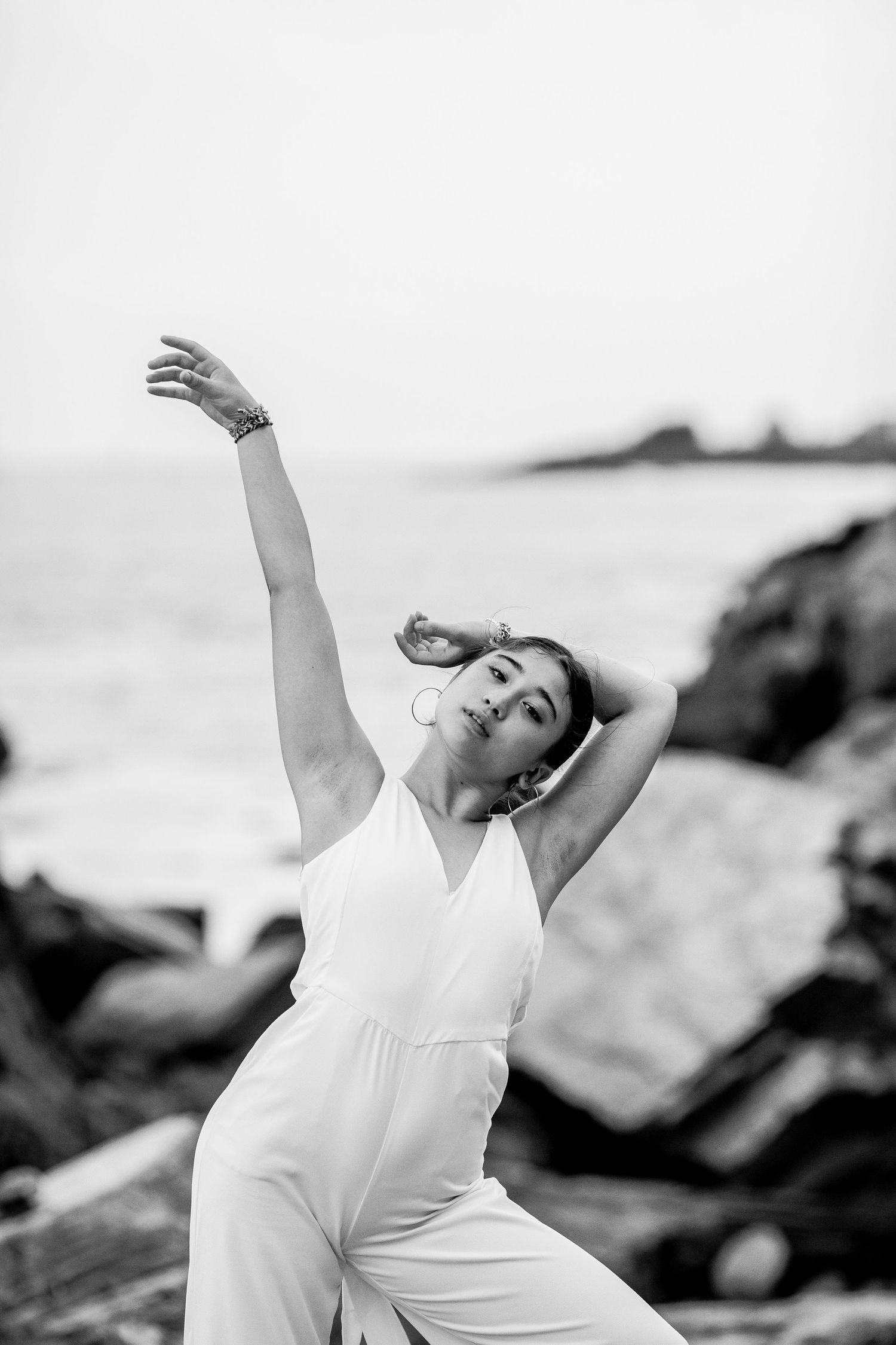 Black and white photo of woman in white jumpsuit dancing expressively on rocky beach with ocean backdrop.