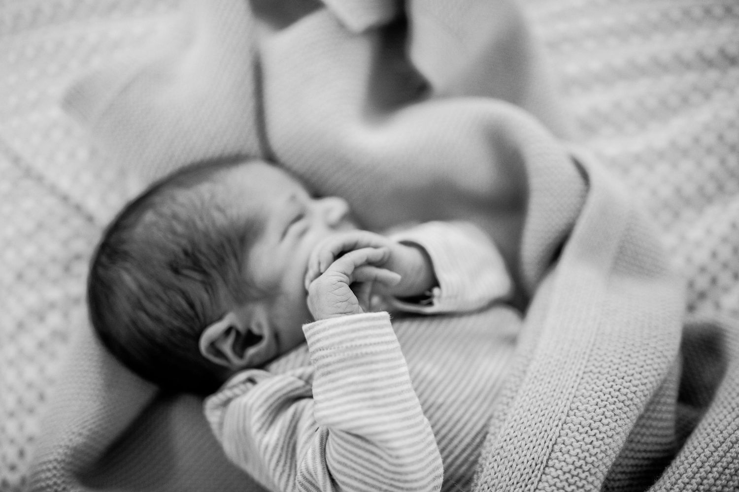 Black and white photo of a sleeping newborn baby wrapped in a striped blanket sucking their fist.