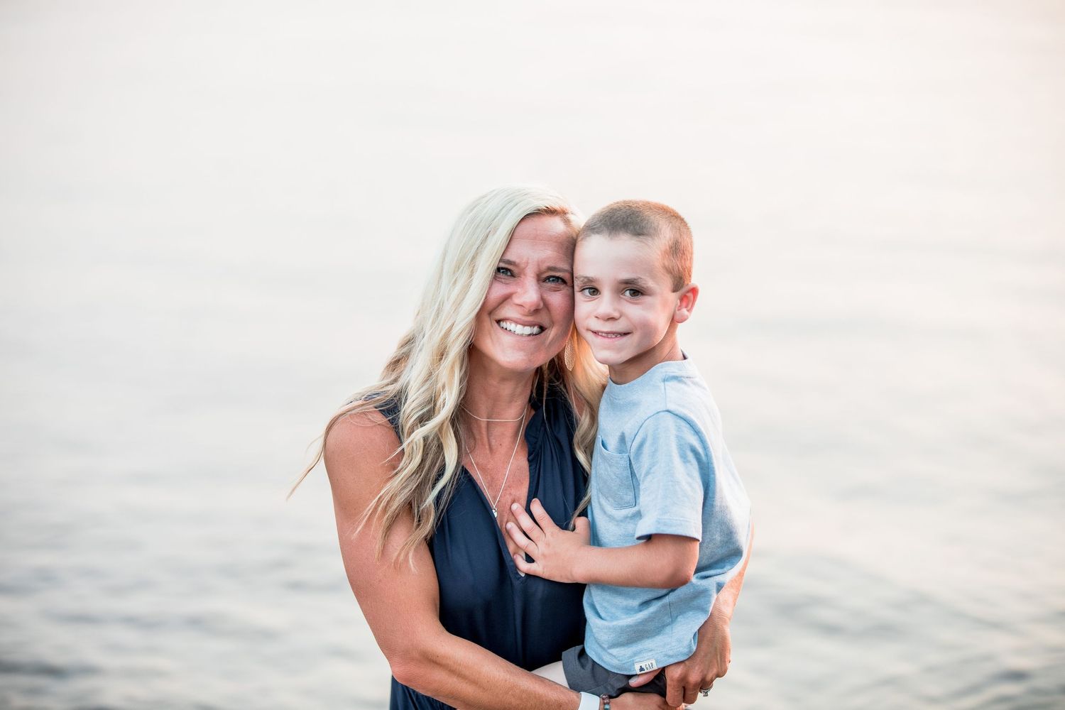 Smiling blonde woman holding young boy near calm water at sunset.