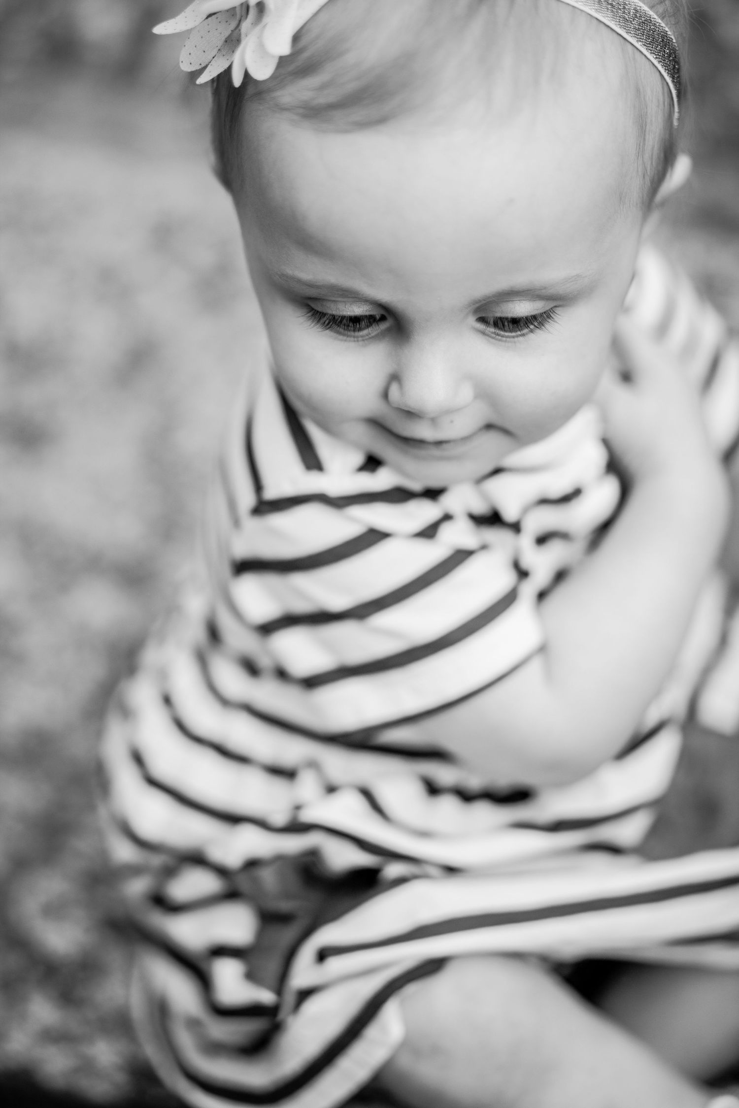 Adorable baby girl in striped dress with flower headband looking down in black and white portrait.