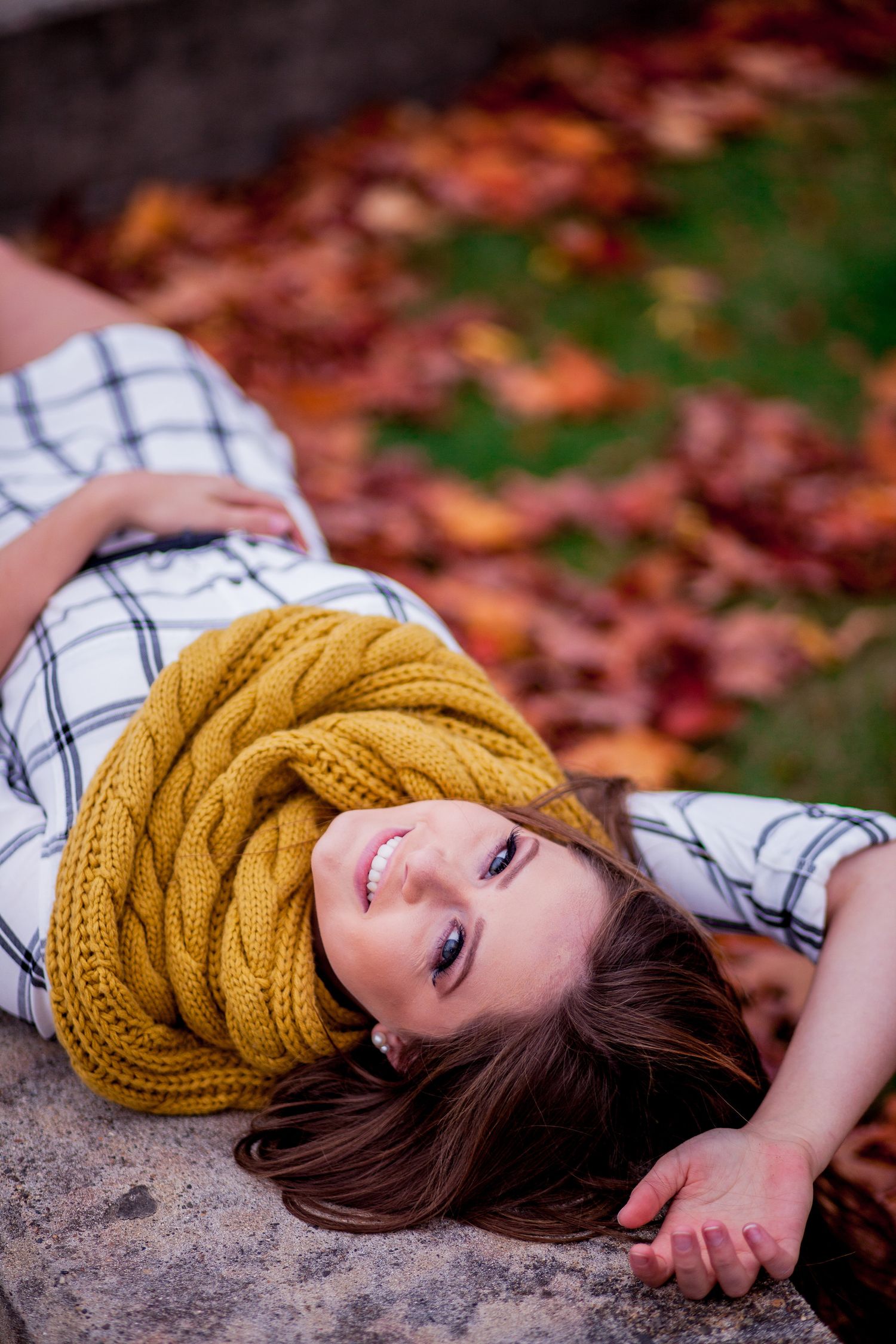 Smiling young woman lying on stone surface wearing yellow knit scarf amid colorful autumn leaves.