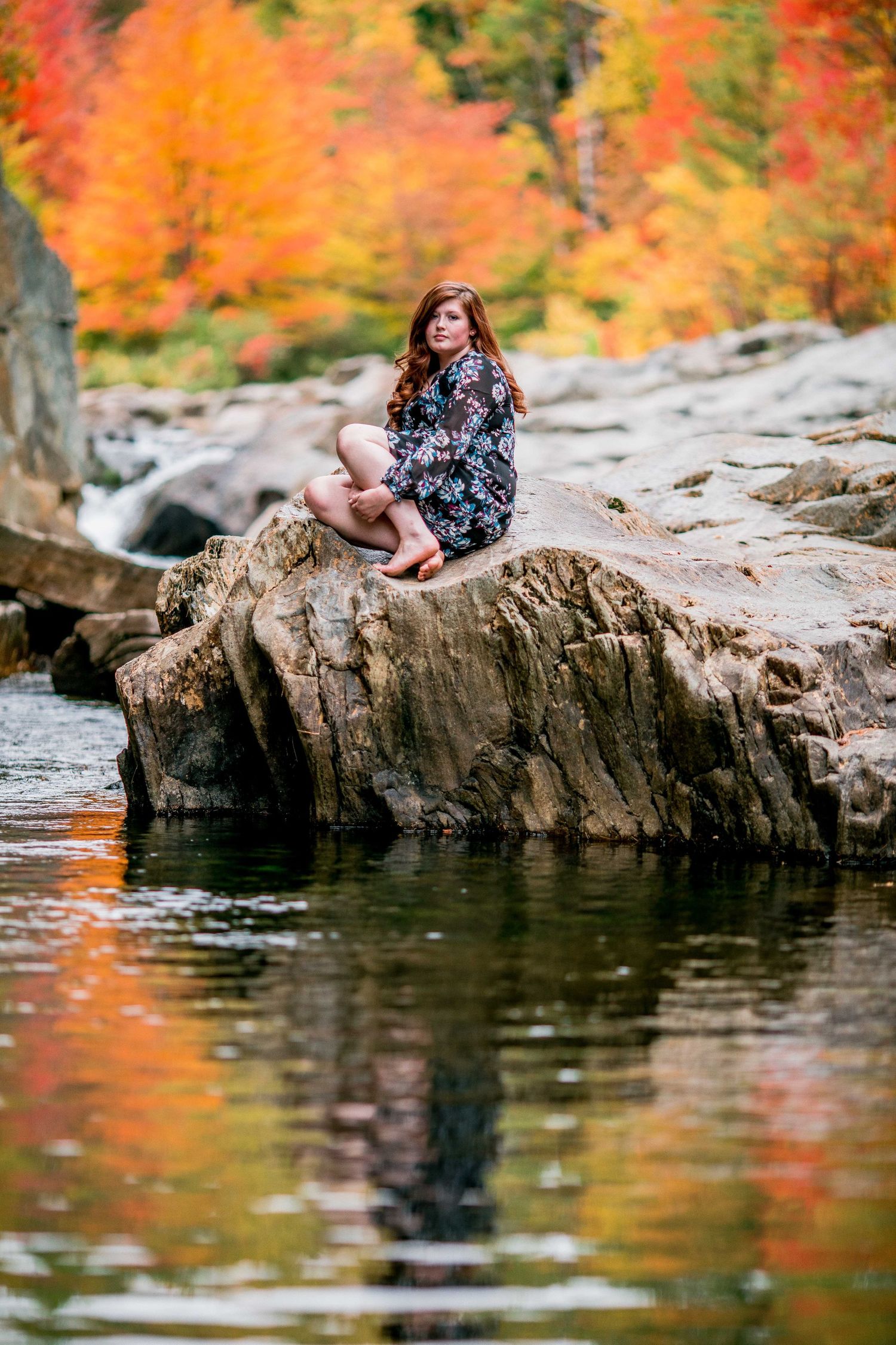 Young woman with red hair sitting cross-legged on rocks by a river with vibrant fall foliage.