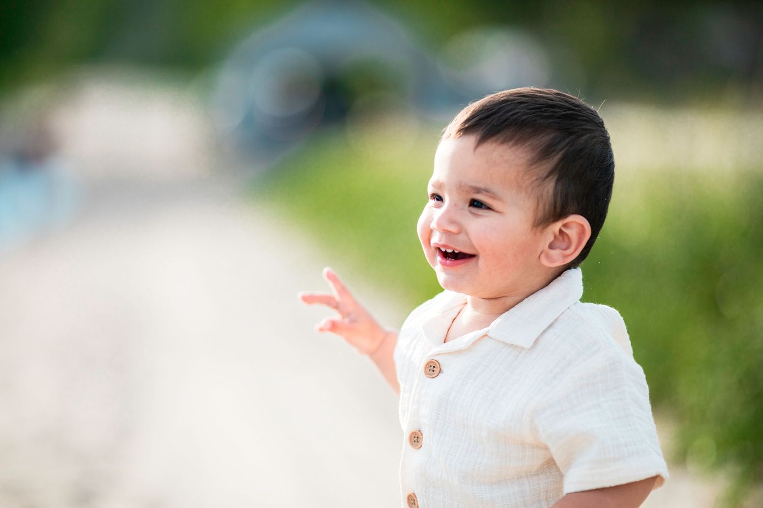 A cheerful toddler boy in a white linen shirt laughs and points outdoors in a sunny green park setting.