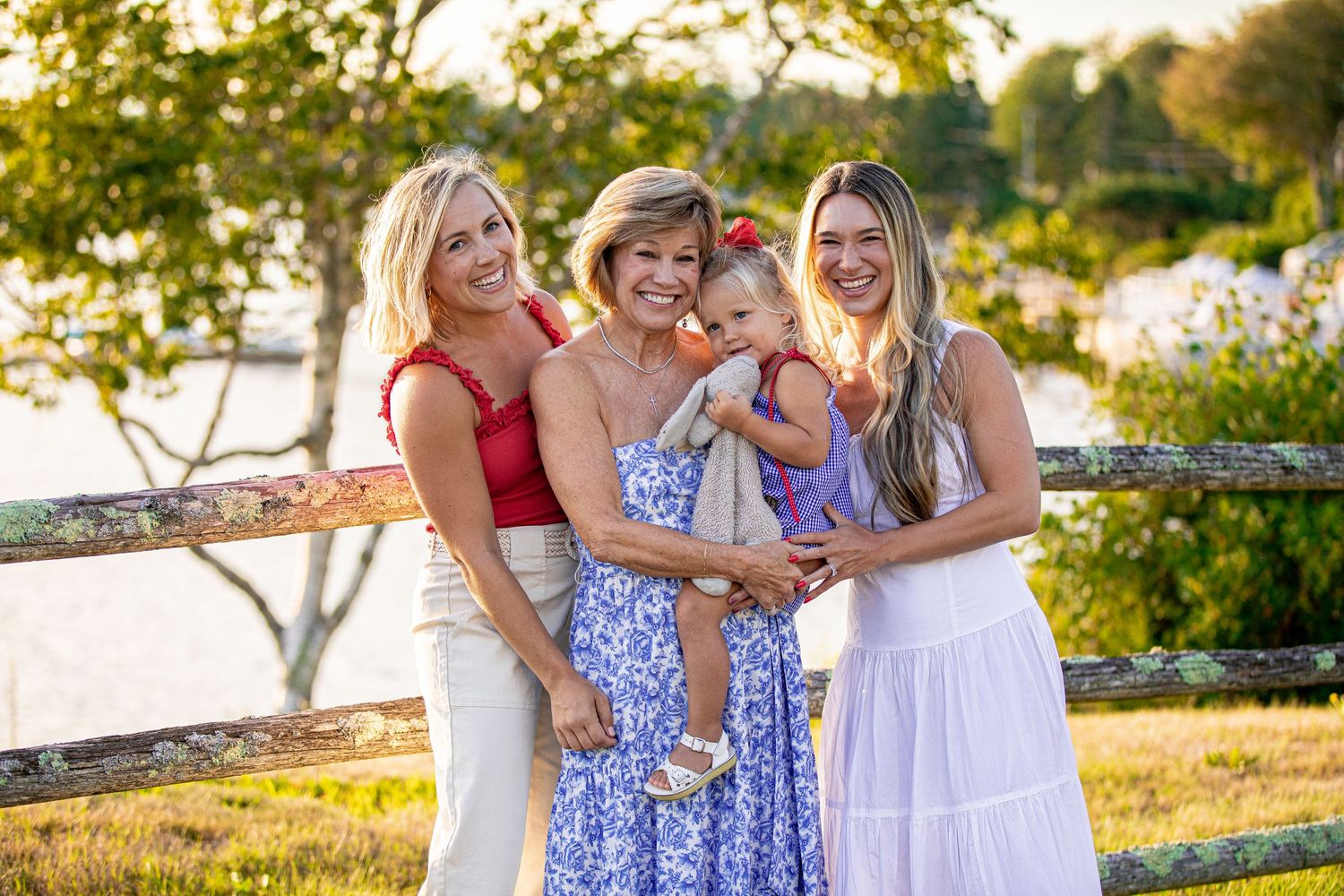 Four generations of women smiling together outdoors on Cape Cod holding baby girl in purple dress.