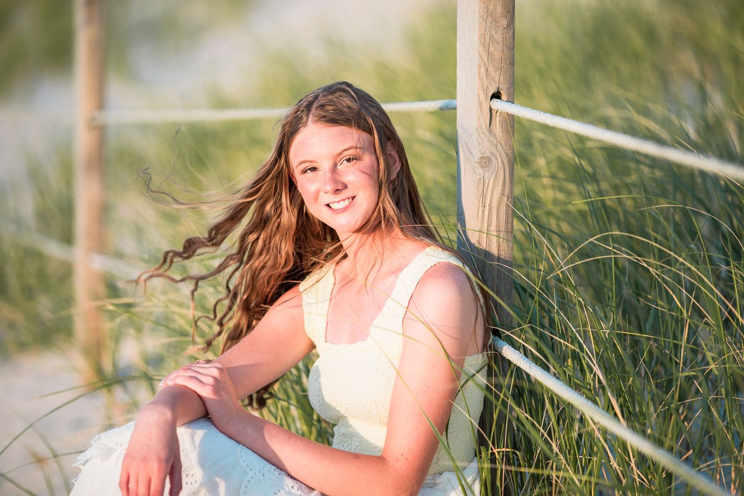 Smiling teenage girl with flowing hair sits by beach grass and wooden fence post on a sunny day.