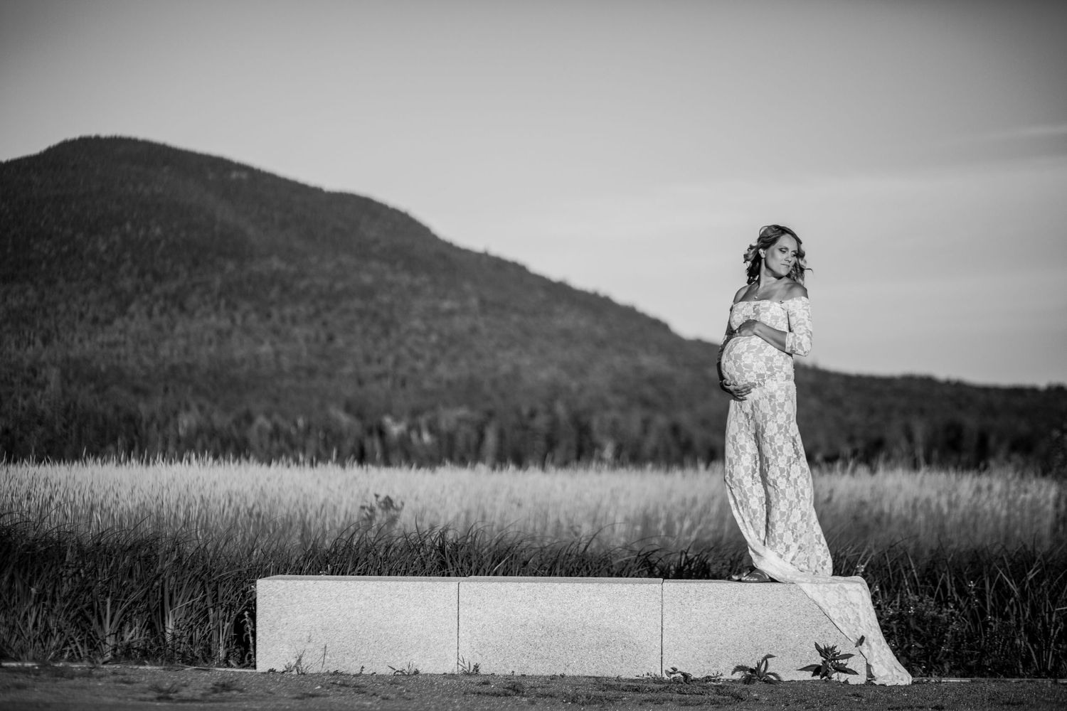 Pregnant woman in white lace gown standing on concrete block in field with mountain backdrop, black and white photo.
