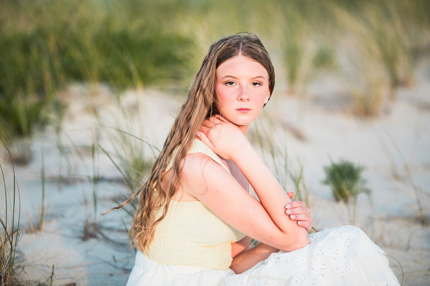 Young girl with long blonde hair in yellow dress sitting on sandy beach dunes with sea grass background.