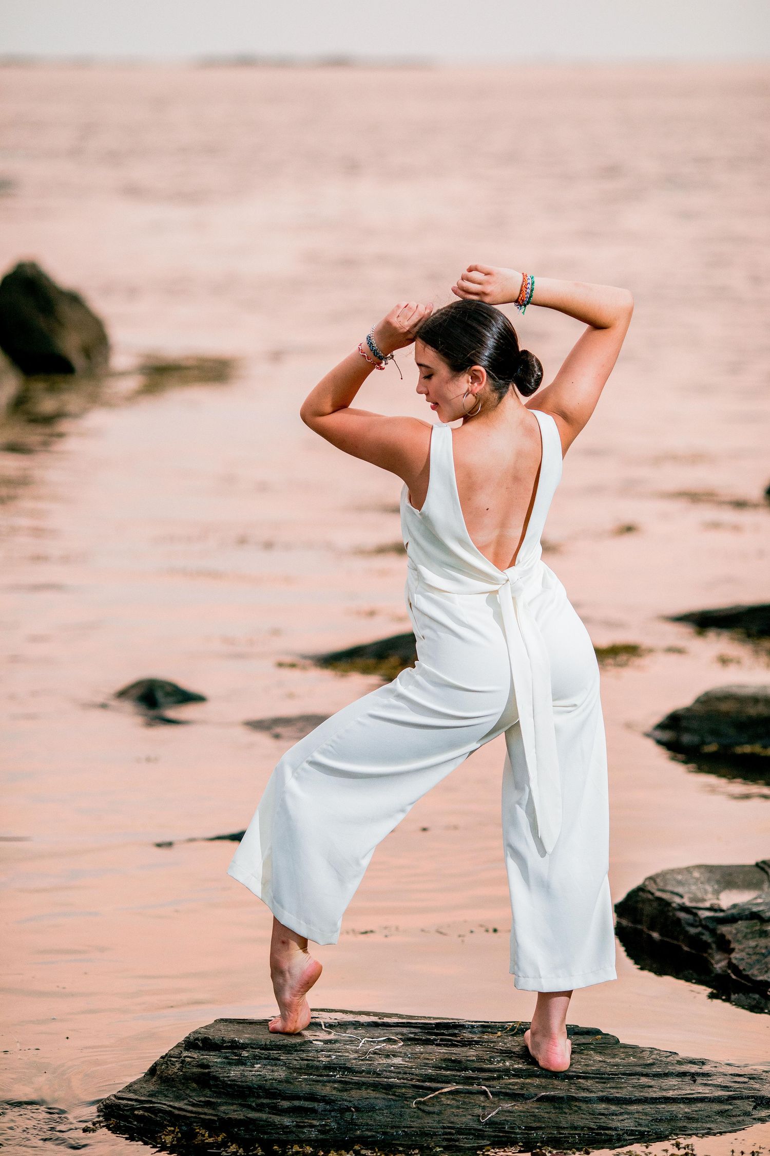 Woman in white backless jumpsuit posing on rocks at beach during golden hour with ocean behind her.