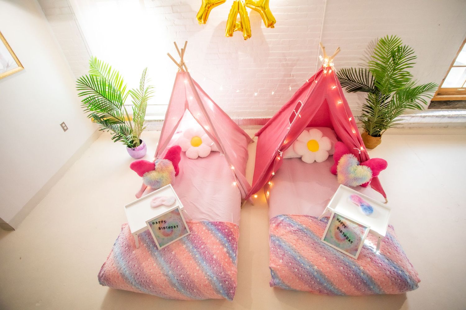 Two pink teepee tents with fairy lights and colorful bedding set up for a kids sleepover party.