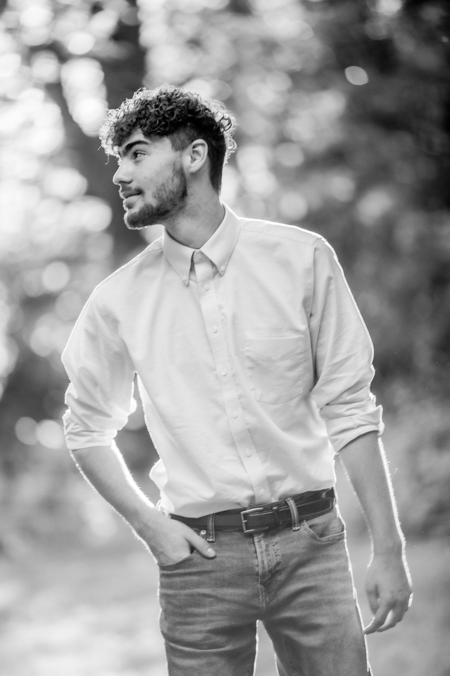 Black and white portrait of a young man with curly hair wearing a white button-up shirt and jeans outdoors.