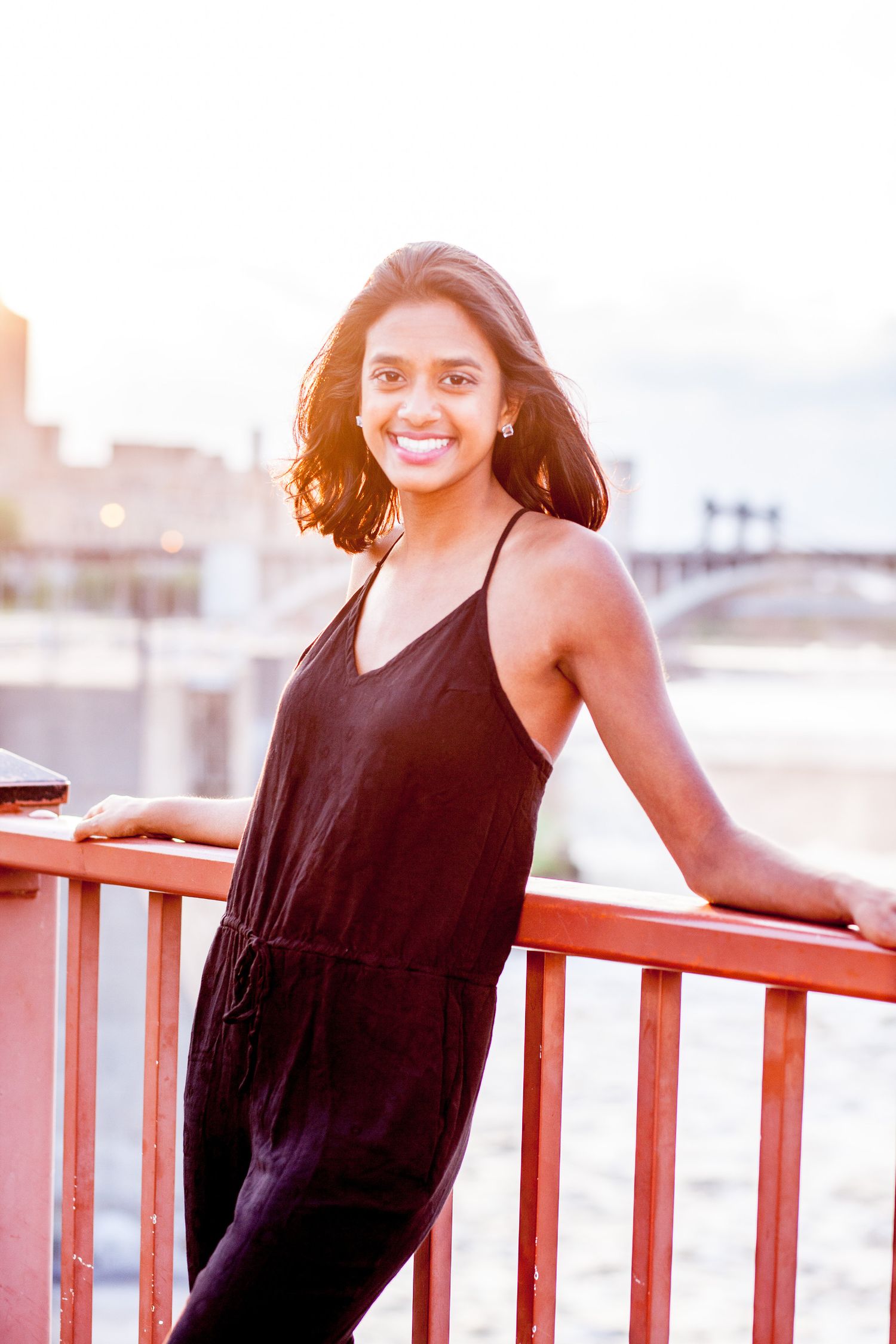 Smiling South Asian woman in black jumpsuit poses on red bridge railing at golden hour sunset.