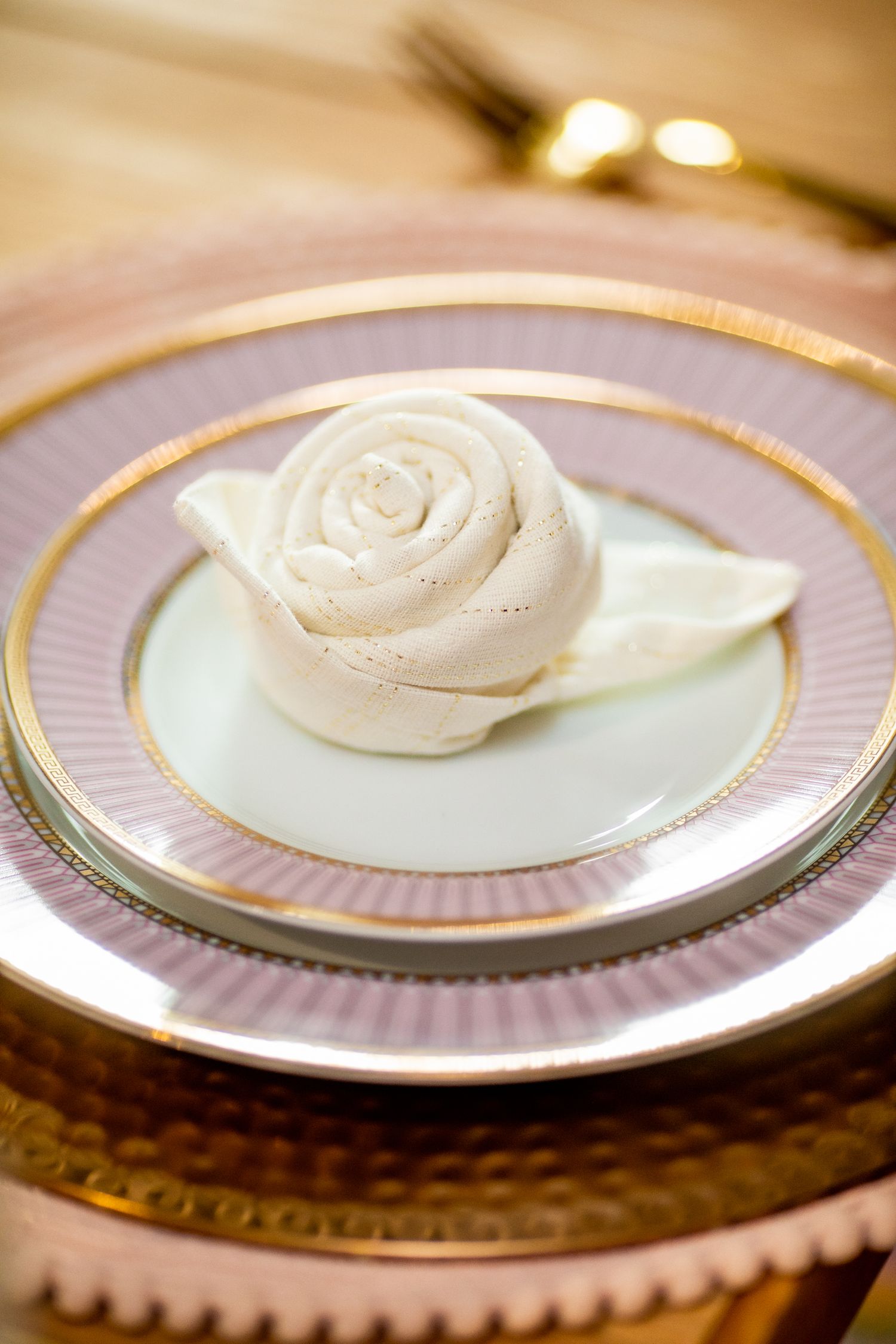 Elegant white napkin folded into a rose shape on stacked gold-rimmed pink and white dinner plates.