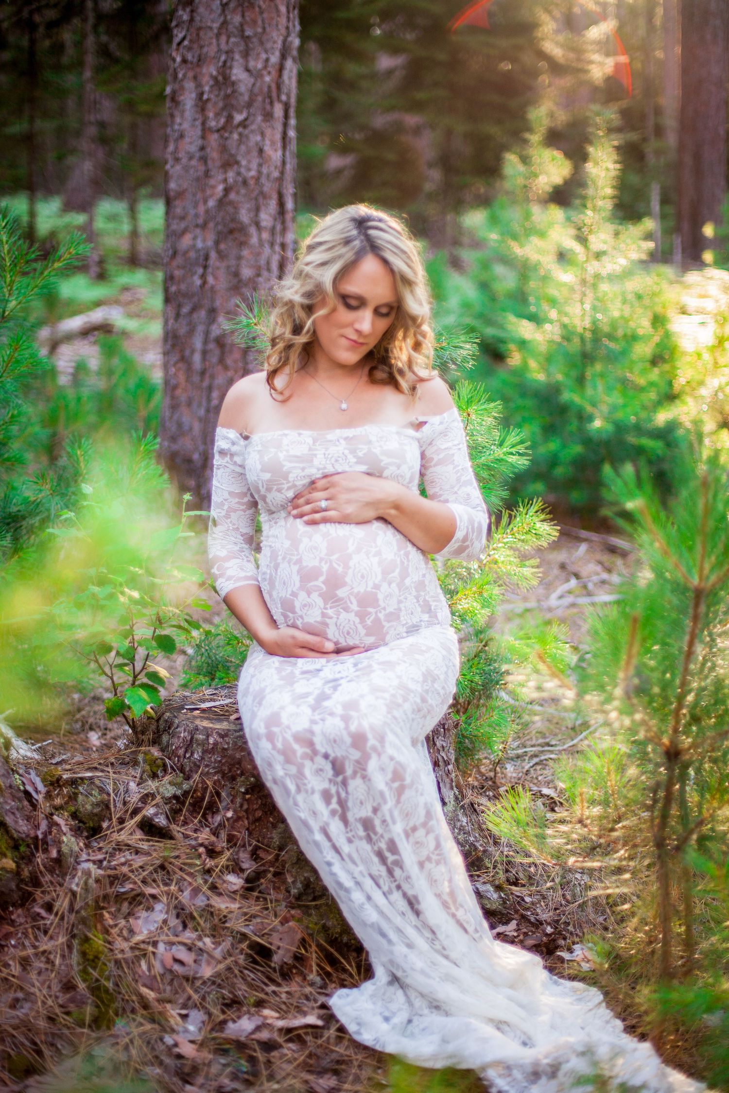 Pregnant woman in white lace gown sitting against tree in sunlit forest, gently holding her baby bump.
