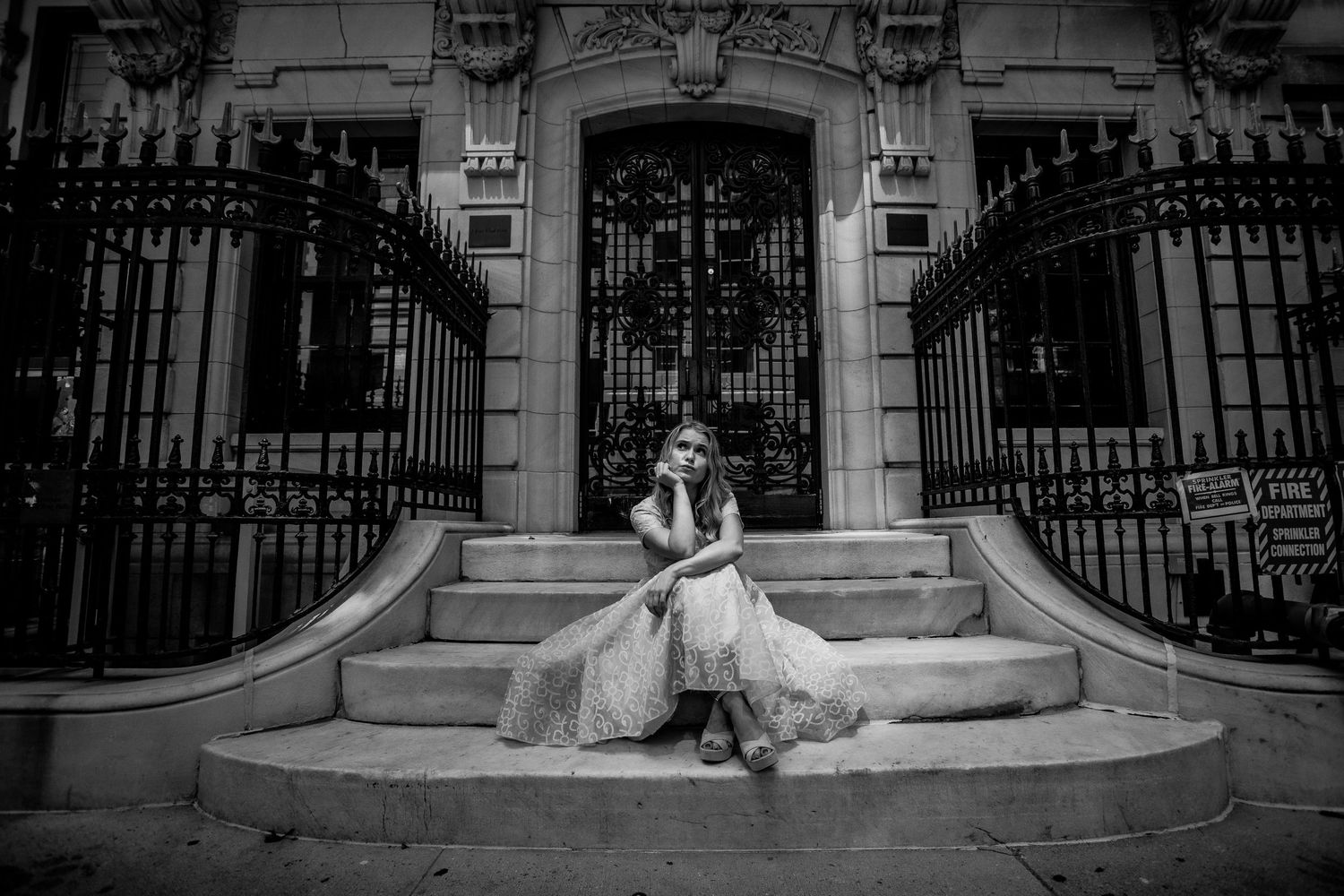 Black and white photo of a young woman in a flowing dress sitting on steps before iron gates.