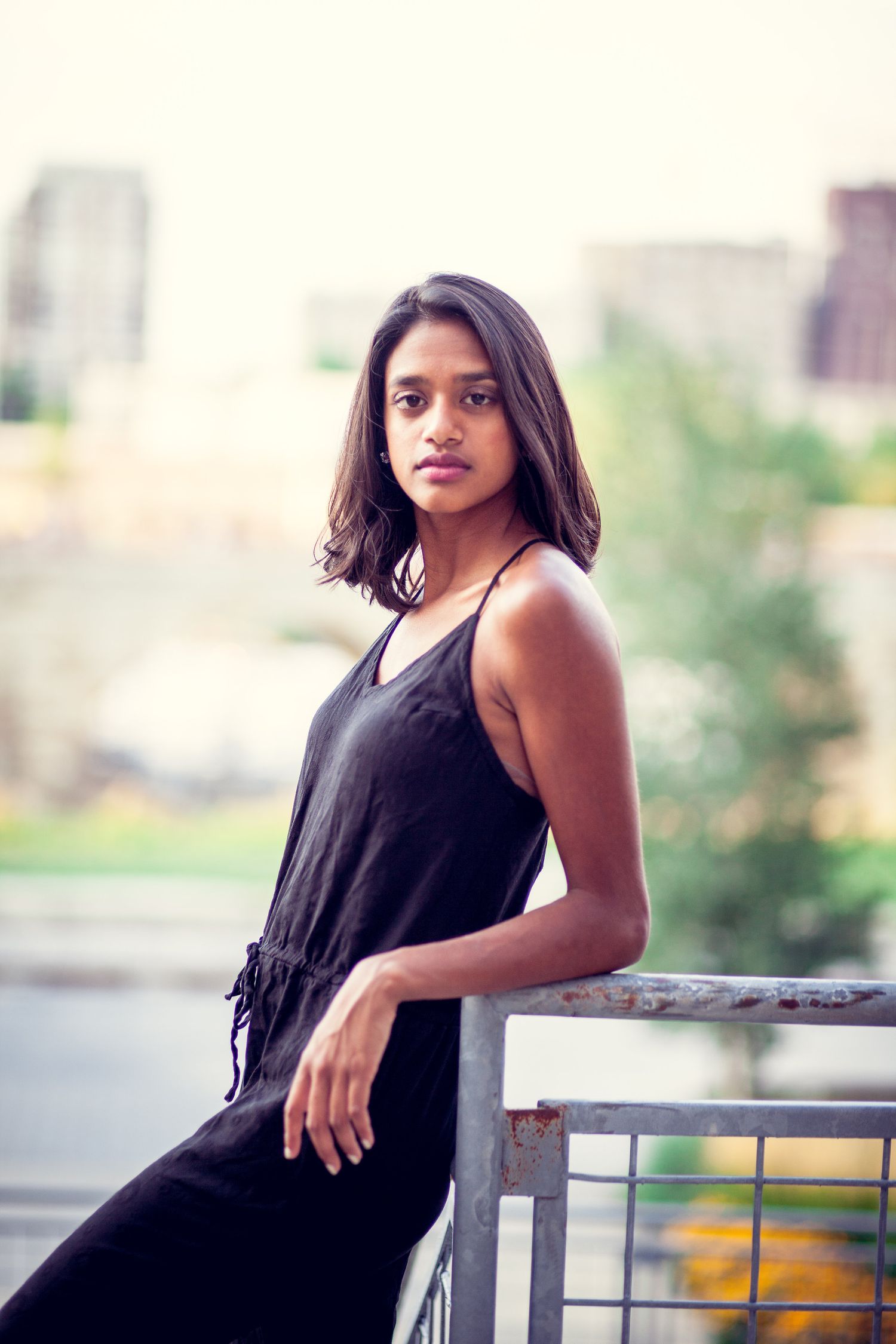 Young South Asian woman in black spaghetti-strap dress leans on metal railing with urban background.