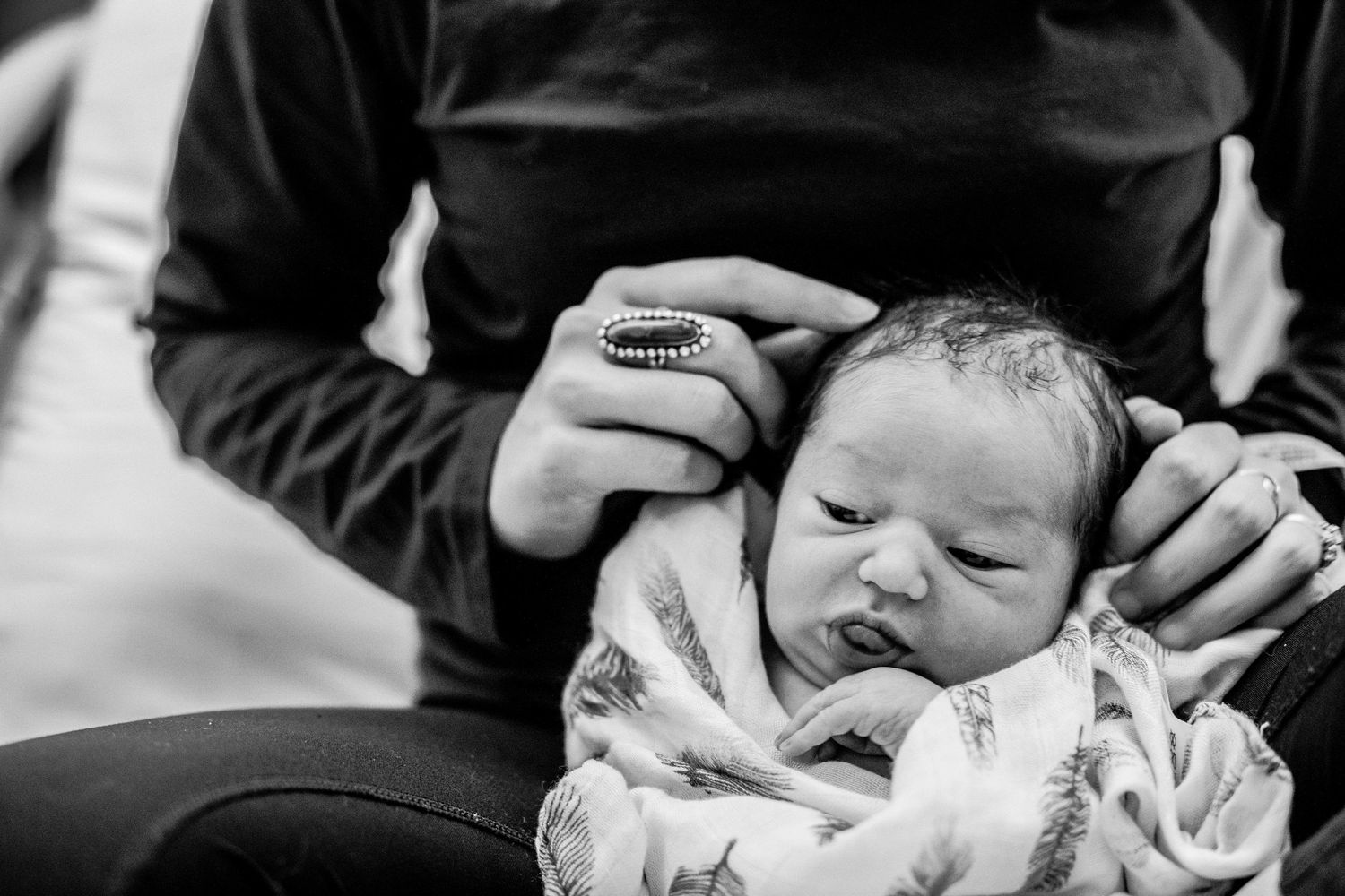 Black and white close-up of newborn baby cradled in parent hands with decorative ring.