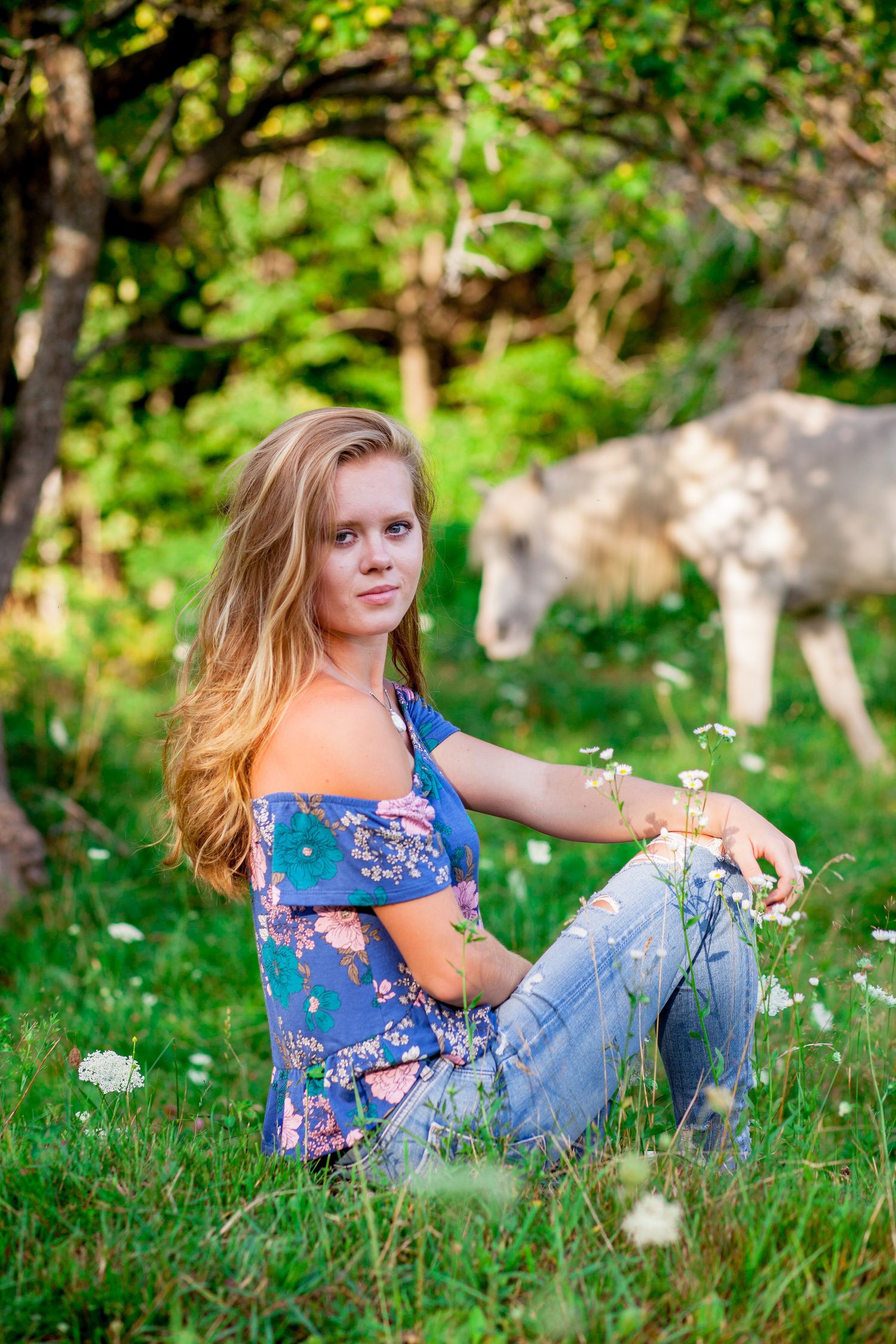 Young blonde woman in floral top and jeans sitting on grass with a white horse grazing in background.