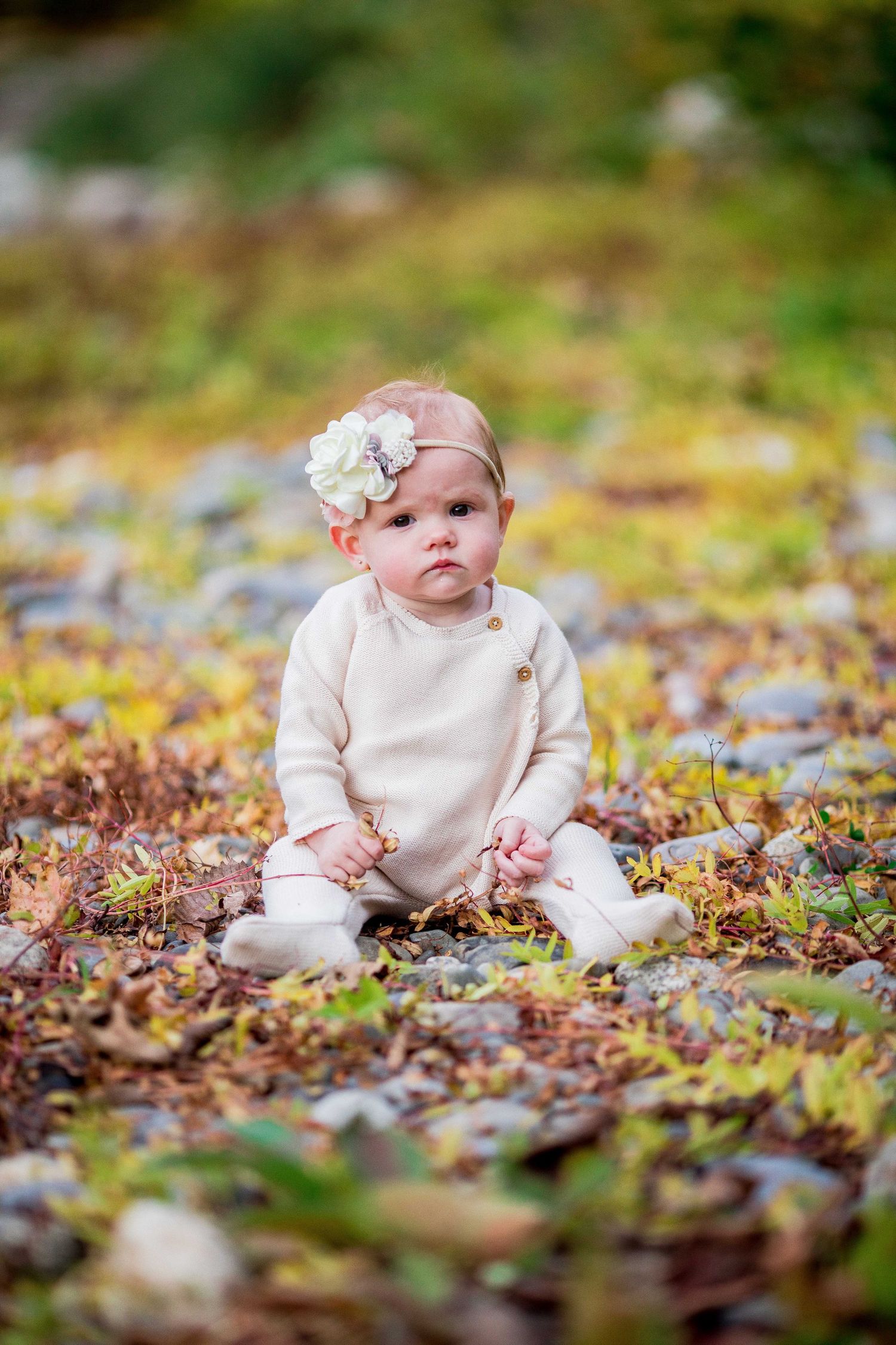 Cute baby girl in cream romper with flower headband sitting on rocky ground covered in fall leaves.