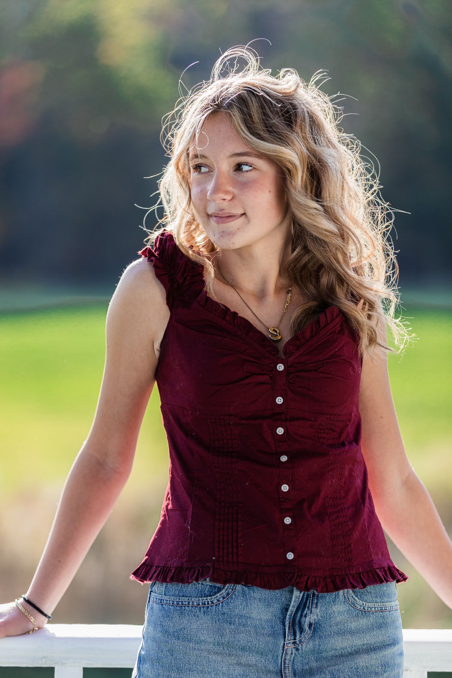 Young blonde teen girl in burgundy top and jeans leaning on white fence outdoors.
