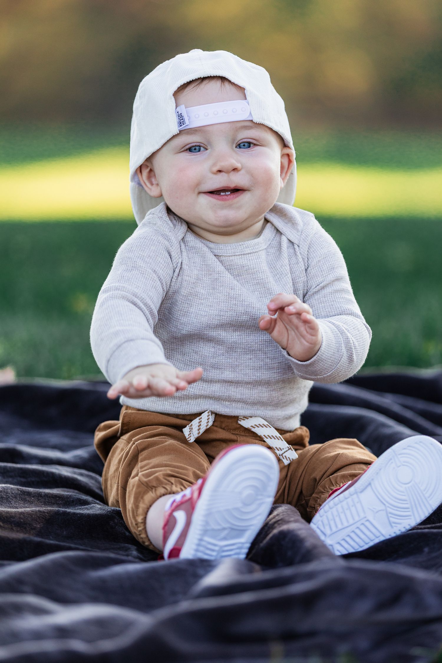 Happy baby boy in white cap and brown pants clapping hands while sitting on blanket in park.