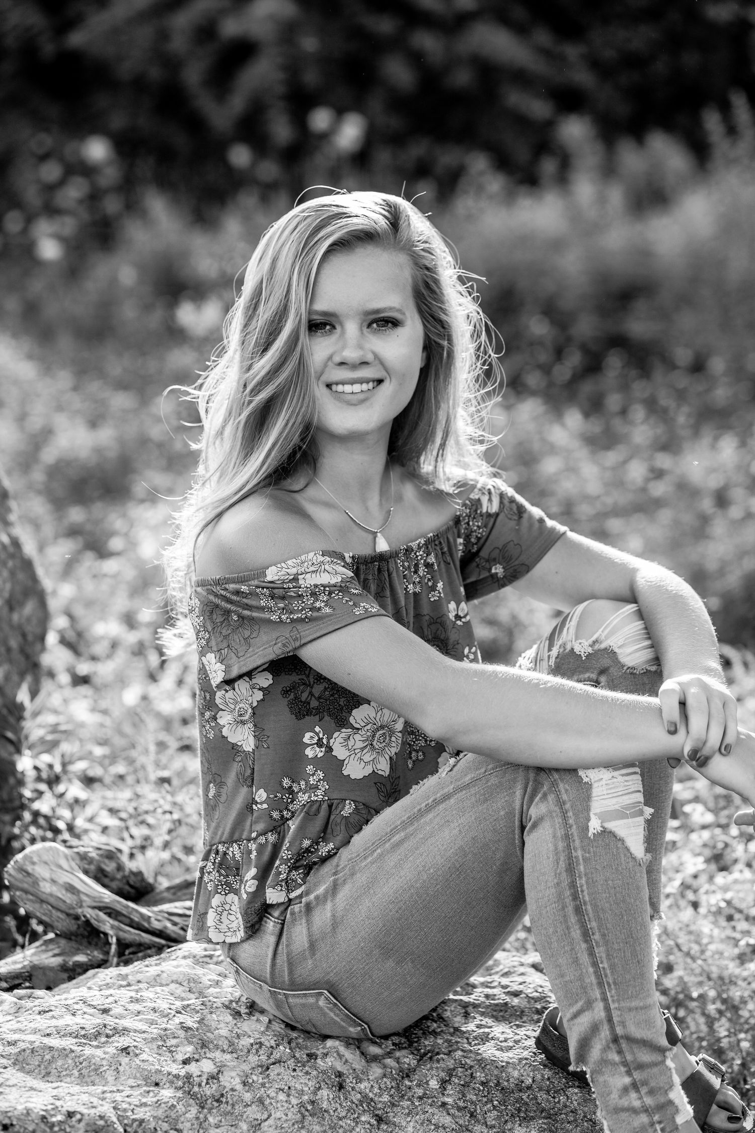 Young blonde woman in floral off-shoulder top and jeans sitting on rocks outdoors, smiling in black and white photo.