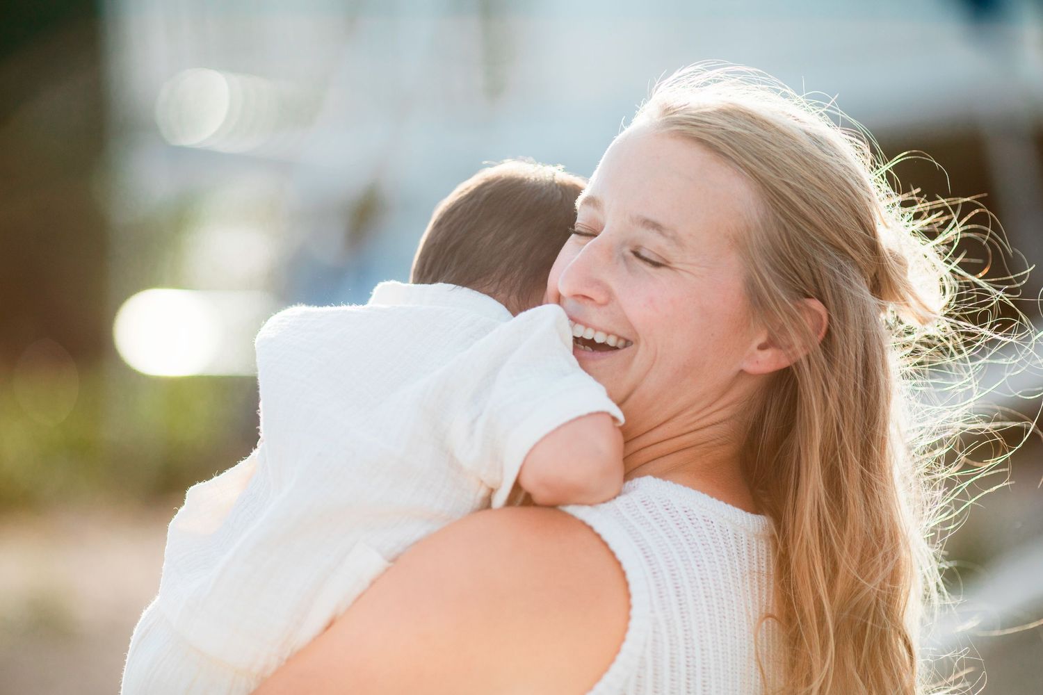Happy young mother with long blonde hair smiling while holding and hugging her baby outdoors in sunlight.